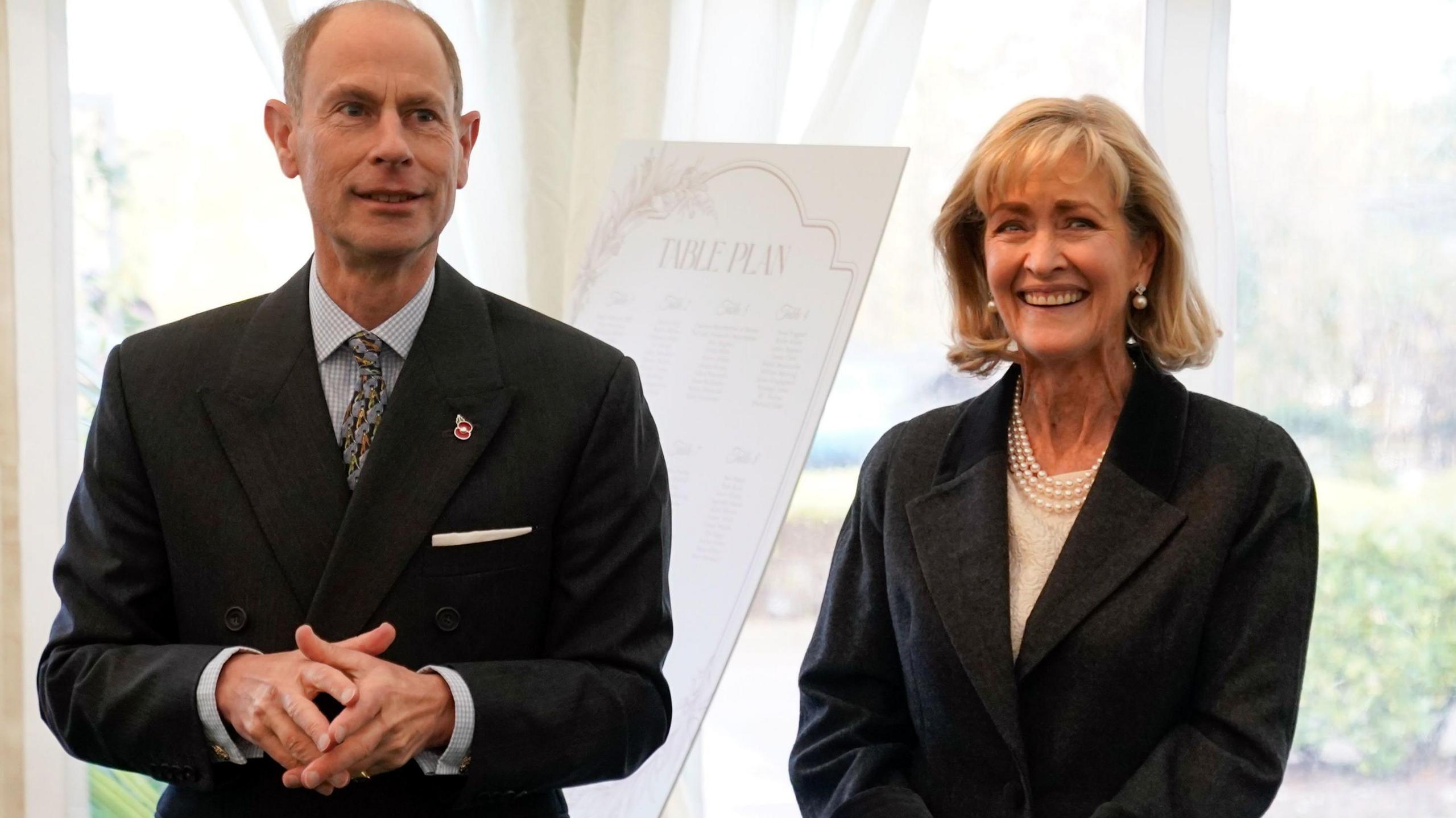 The Duke of Edinburgh, Prince Edward is smiling at the camera and wearing a black suit, blue shirt and patterned tie. Penelope Knatchbull the Countess Mountbatten of Burma is smiling at the camera, is wearing a black jacket, over a cream jumper with three strings of pearls round her neck.