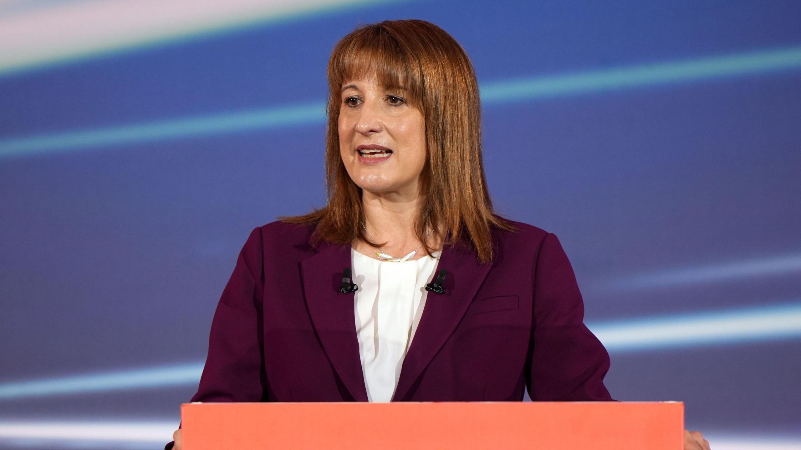 Chancellor Rachel Reeves wears a plum-coloured suit and white shirt as she stands in front of a red podium.