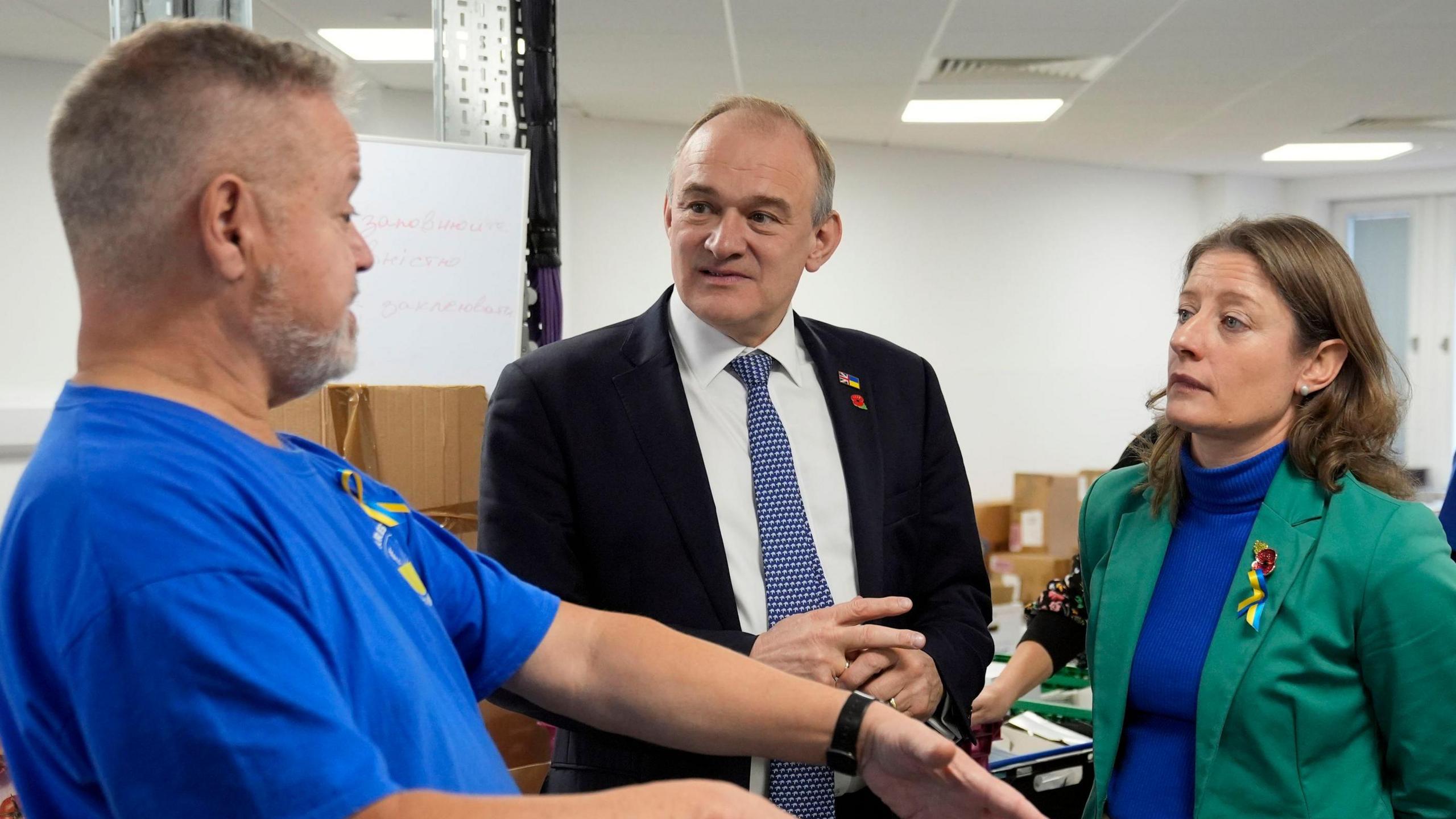 Sir Ed Davey in a dark suit and blue and white tie looking at a man with short white hair and a beard while he is talking and demonstrating something with his arms. MP Helen Maguire is standing to the right in a blue turtle-neck and green blazer looking at the man talking.