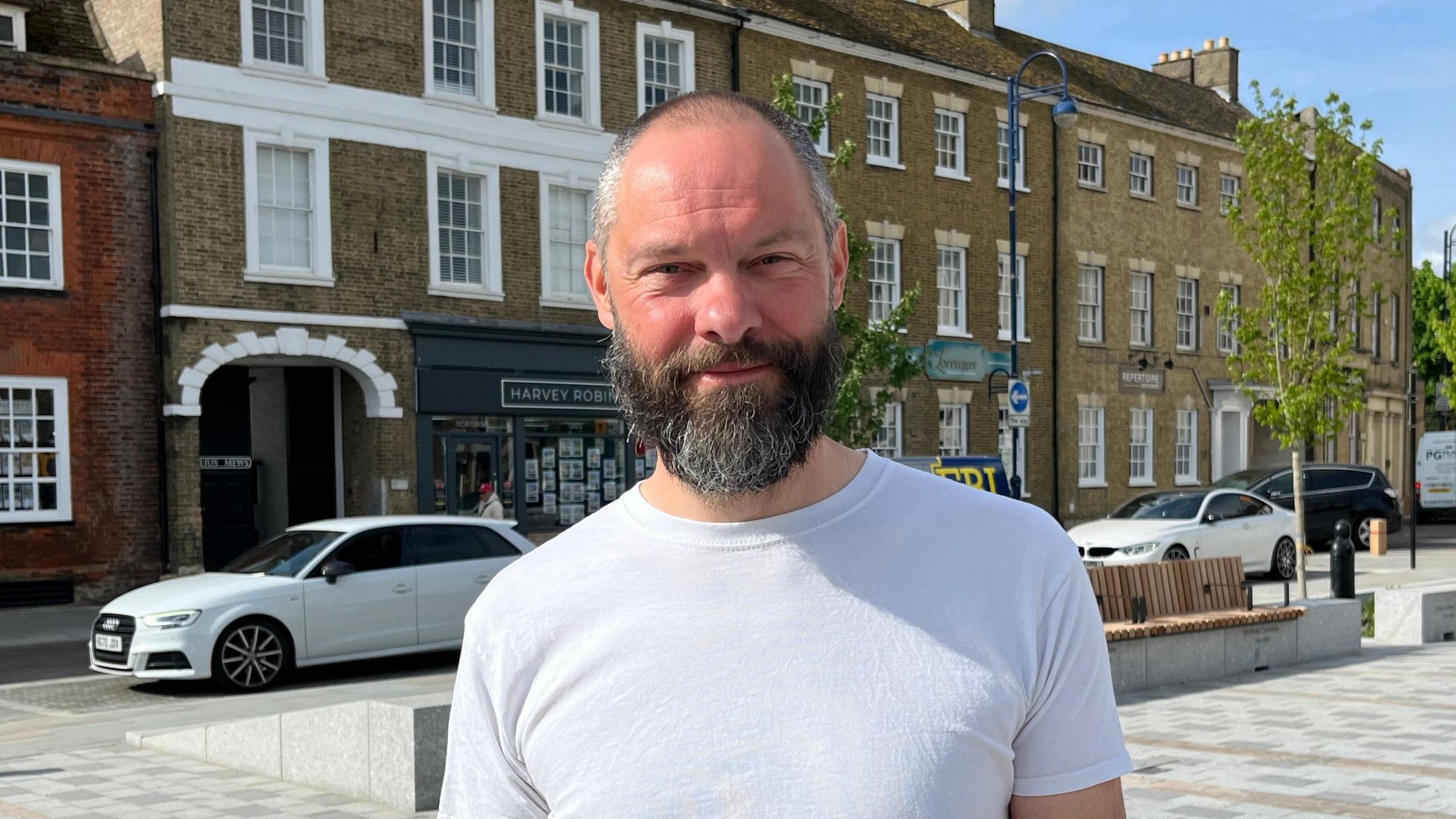 Richard Slade is standing in the square while wearing a white top. He is smiling at the camera and has short hair and a beard, both are a mix of brown with some grey. Behind him is some seating on the square and trees and three-storey buildings