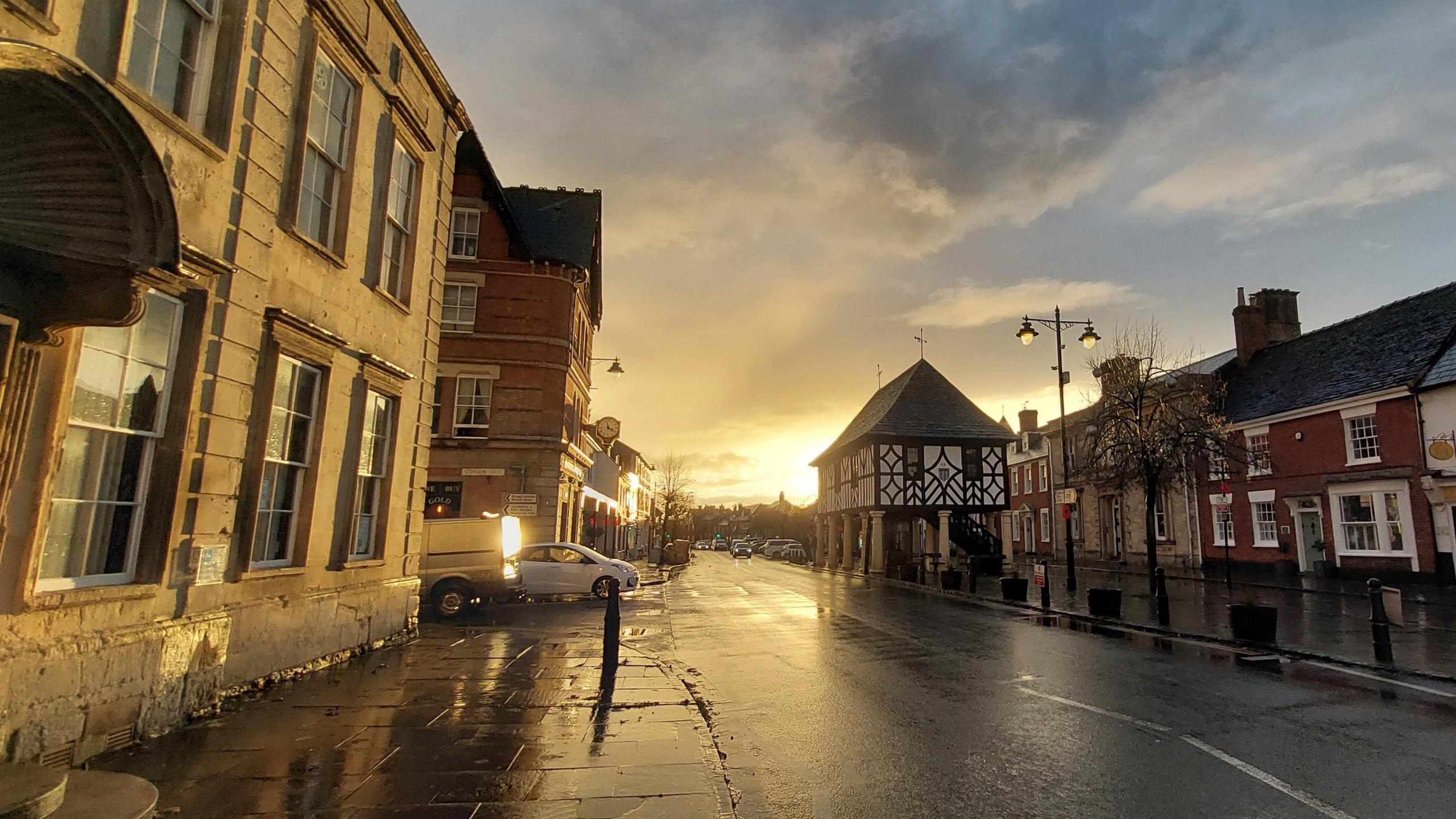 A high street with old style buildings and few cars on the road. The sun is setting and there is a golden colour to the street, with some patches of puddles from recent rainfall.