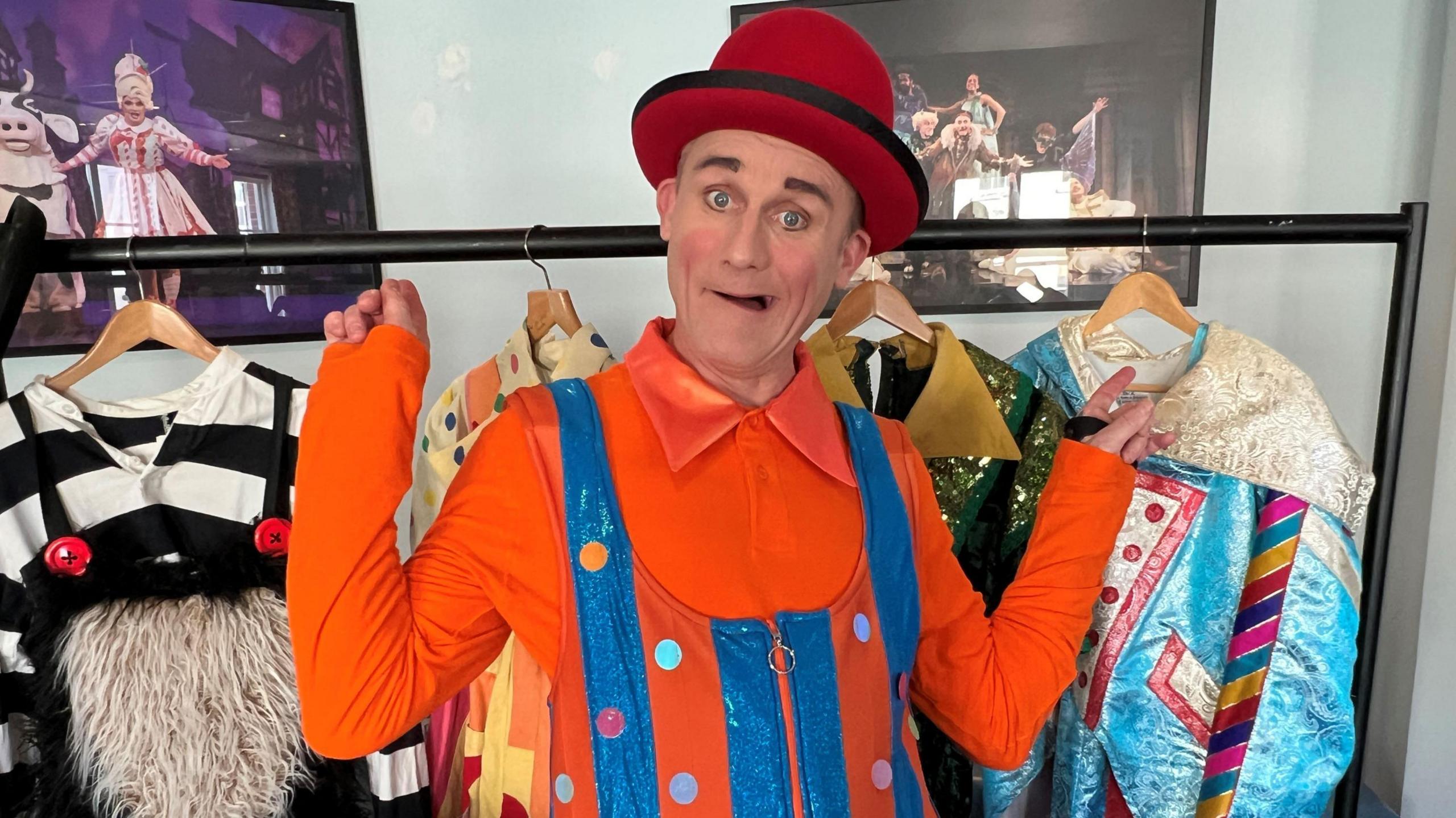 A man dressed as a clown, with a red hat and orange shirt, stands in front of a rack of different outfits. His name is Tweedy the clown and he is appearing in Aladdin at the Everyman Theatre in Cheltenham