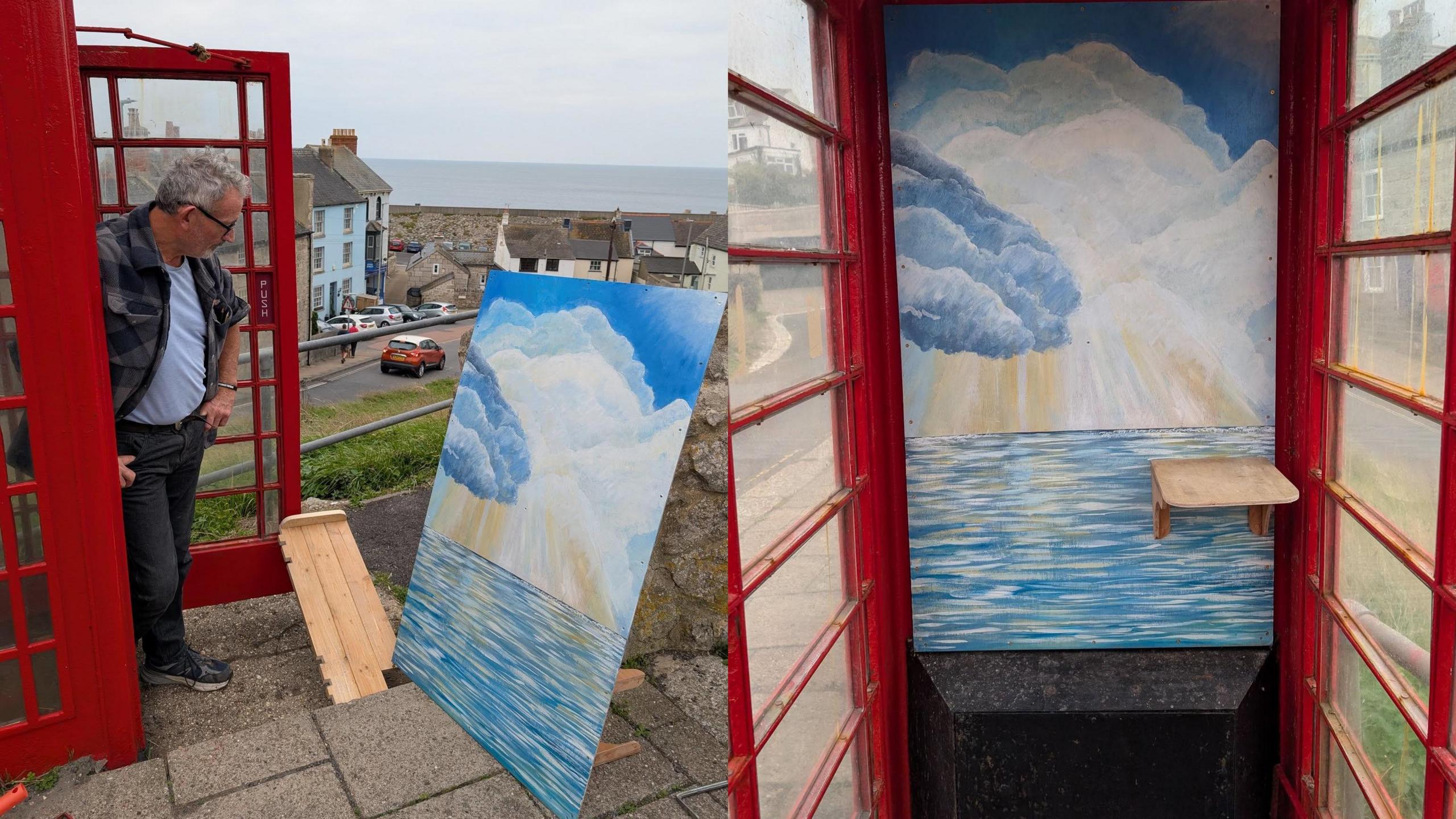 A red phone box with a man stood looking at a paiting with blue sea and white clouds.