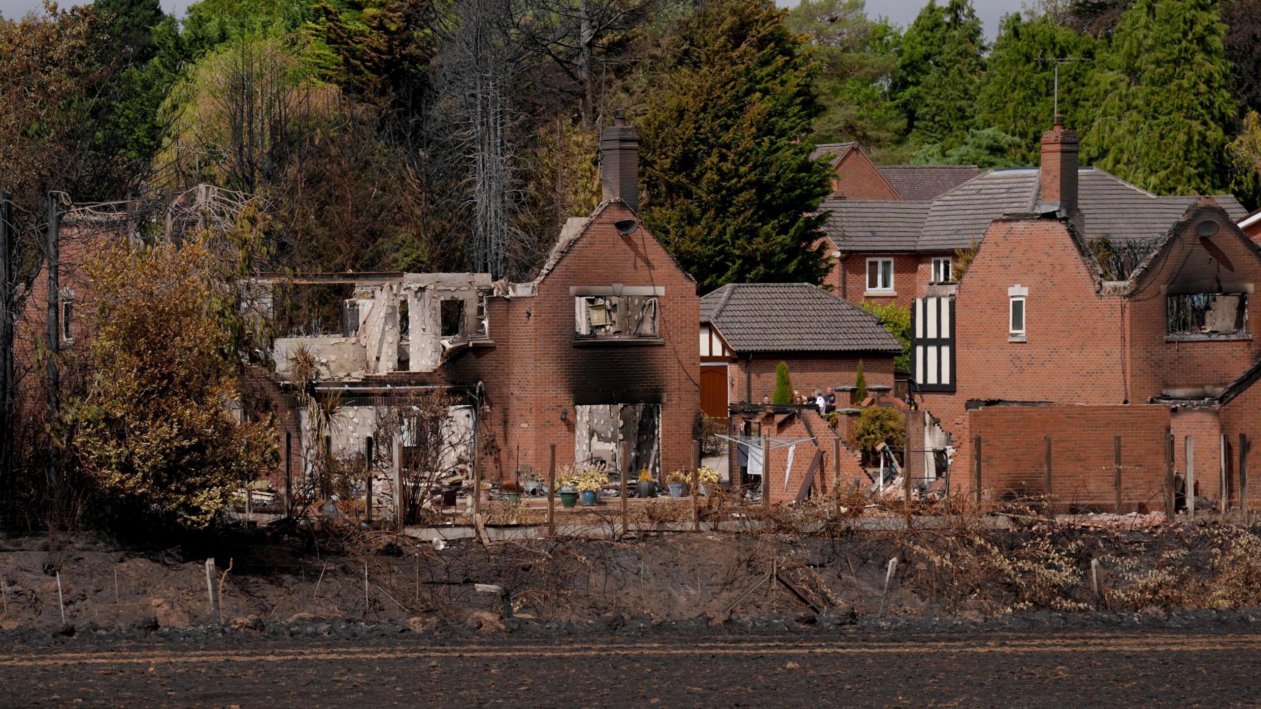 A distant photo of several houses completely destroyed by fire. They do not have roofs and some walls have fallen away. they are blackened with smoke 