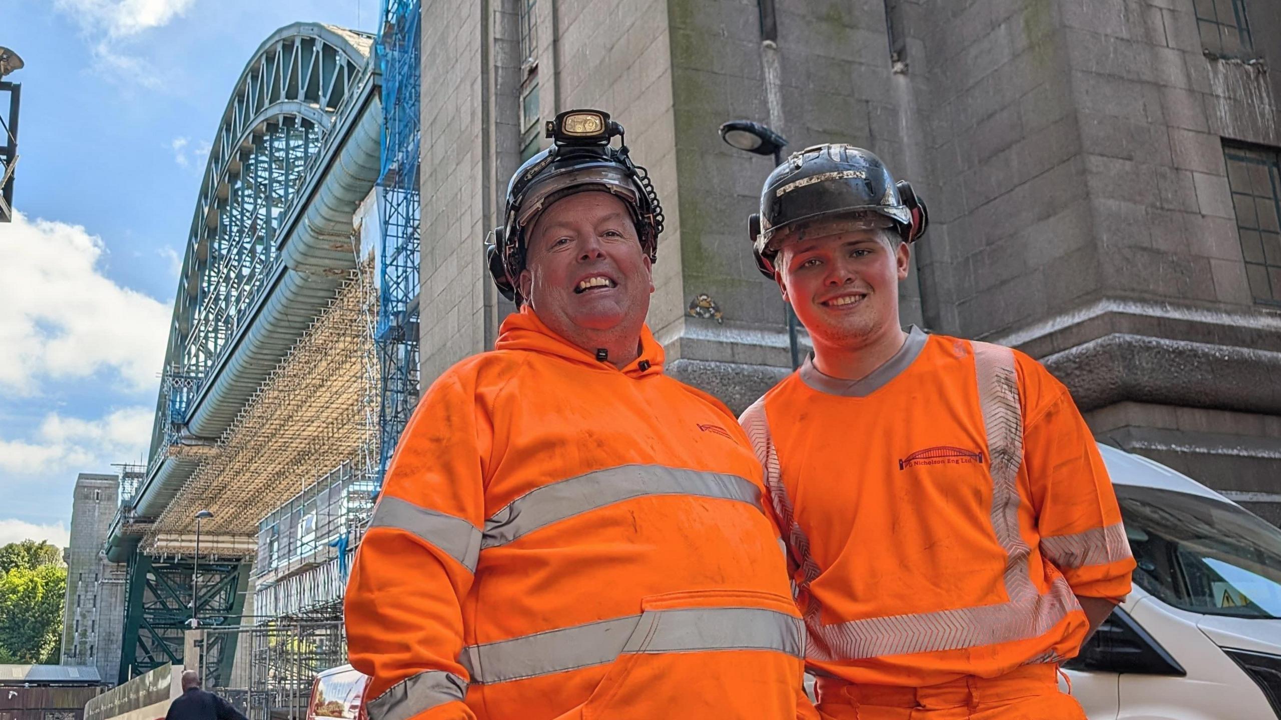 Paul and Charlie stand smiling with the dominating Tyne Bridge towering behind them in the background. 