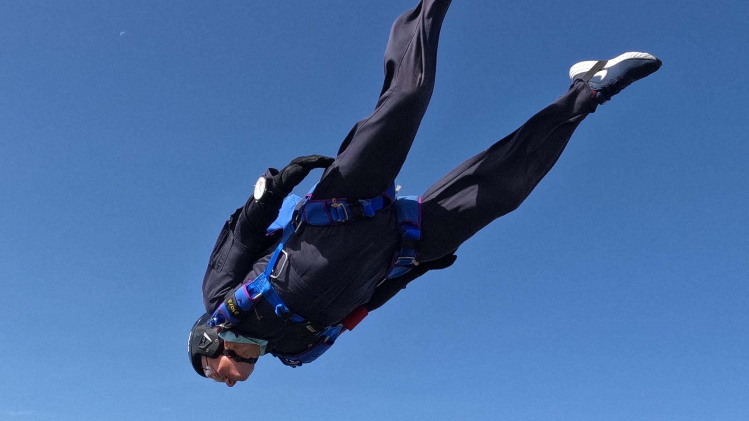 Chris Copeland in freefall against a bright blue sky. He is head down and legs up, wearing a skydiving suit, blue parachute harness, a helmet and protective goggles. 