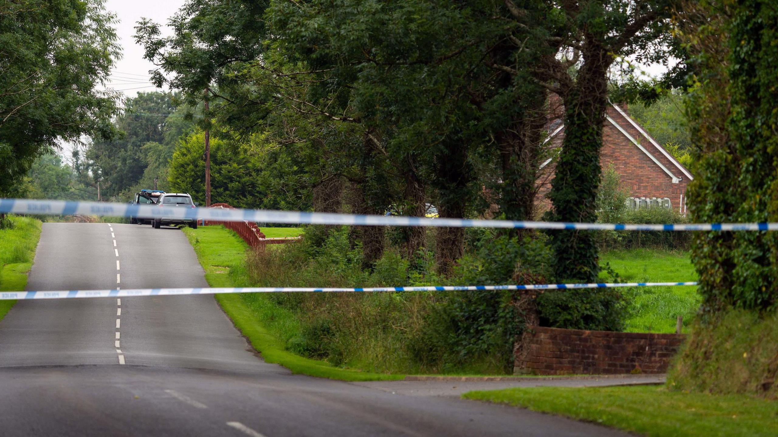 A wide shot of a blue and white police cordon, stretching across a rural road outside Maguiresbridge village. The side of a large redbrick house is partially visible, but the view is blocked by a line of tall trees. Two vehicles are parked at the top of a hill, outside a wooden fence which surrounds the house.