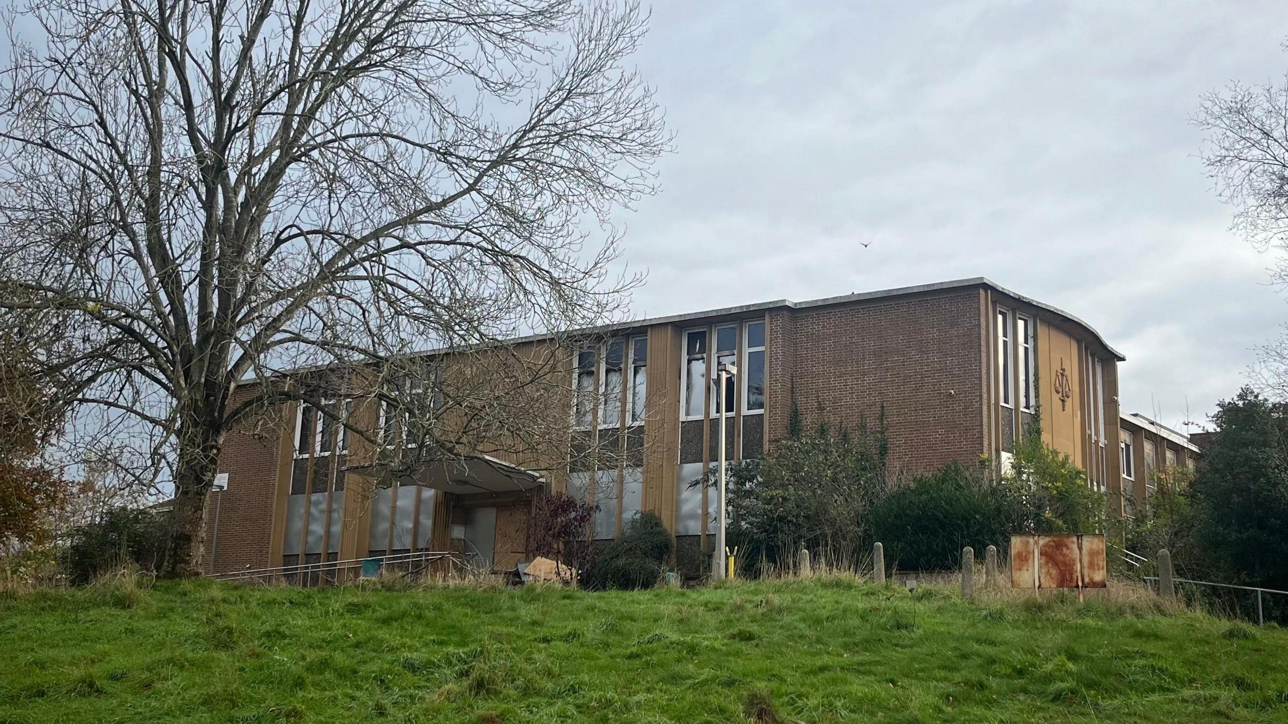A building at the top of a mildly sloped grass bank. The brick building is large with windows. There are a number of windows.