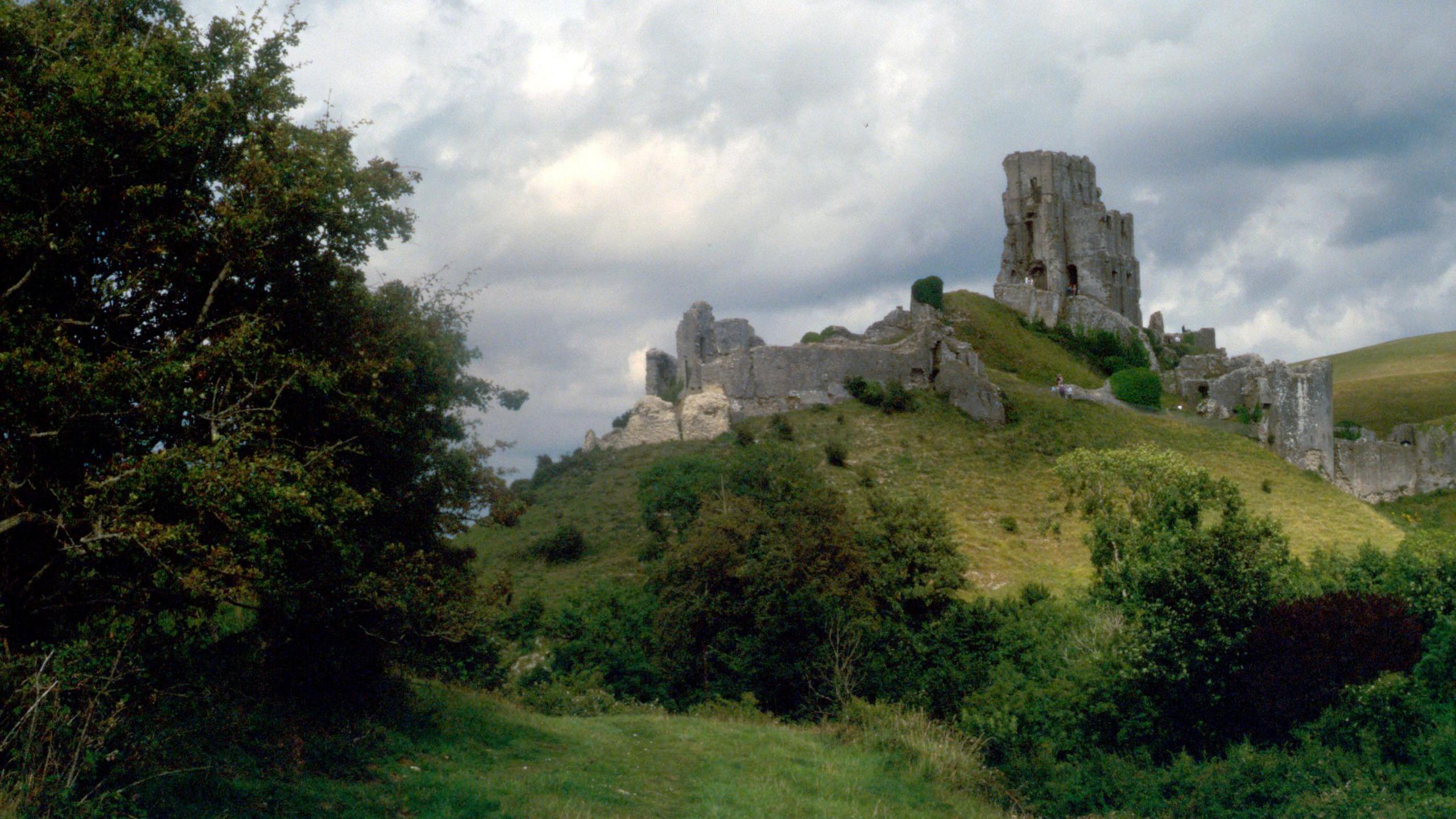 View looking up at Corfe Castle ruins which sits on top of a large mound surrounded by trees