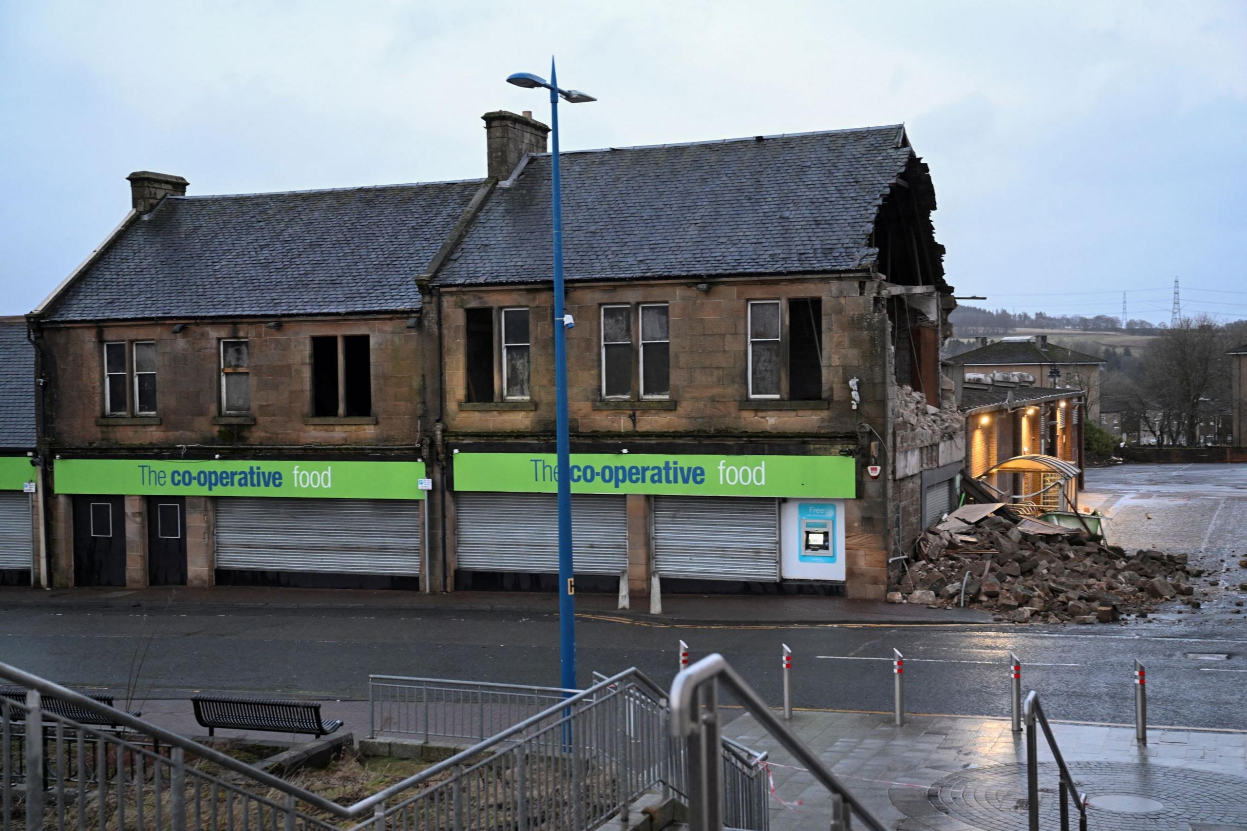 The Co-Op in Denny near Falkirk viewed from the front is missing a whole stone wall on the right. The rubble is strewn across the road. 