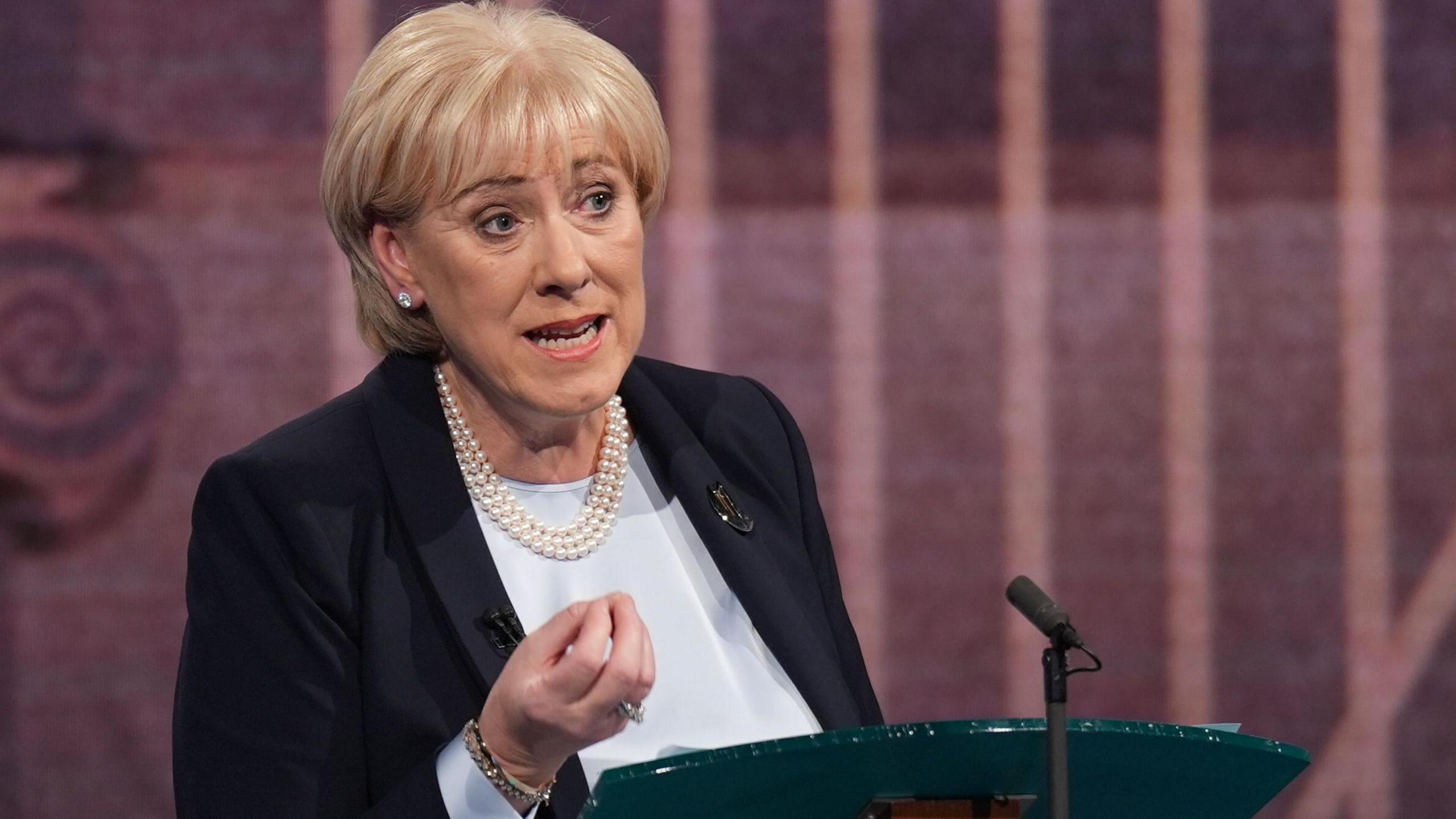 Heather Humphreys wearing a black suit and white top with a pearl necklace. She has blonde hair. She is standing at a lecturn.