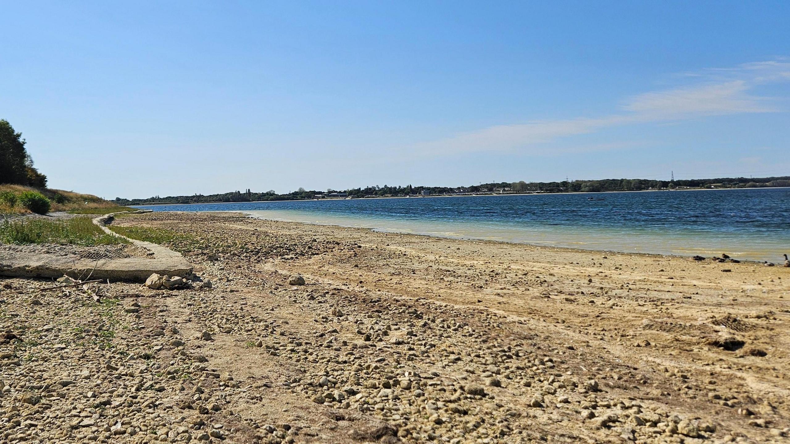 A view of Grafham Water lake and reservoir from the perspective of someone standing on the shore. It shows low water levels as the shore is widely exposed with many stones on . The water is turquoise then a deep blue, and there are pylons in the distance. To the left is a grassy bank and trees. 
