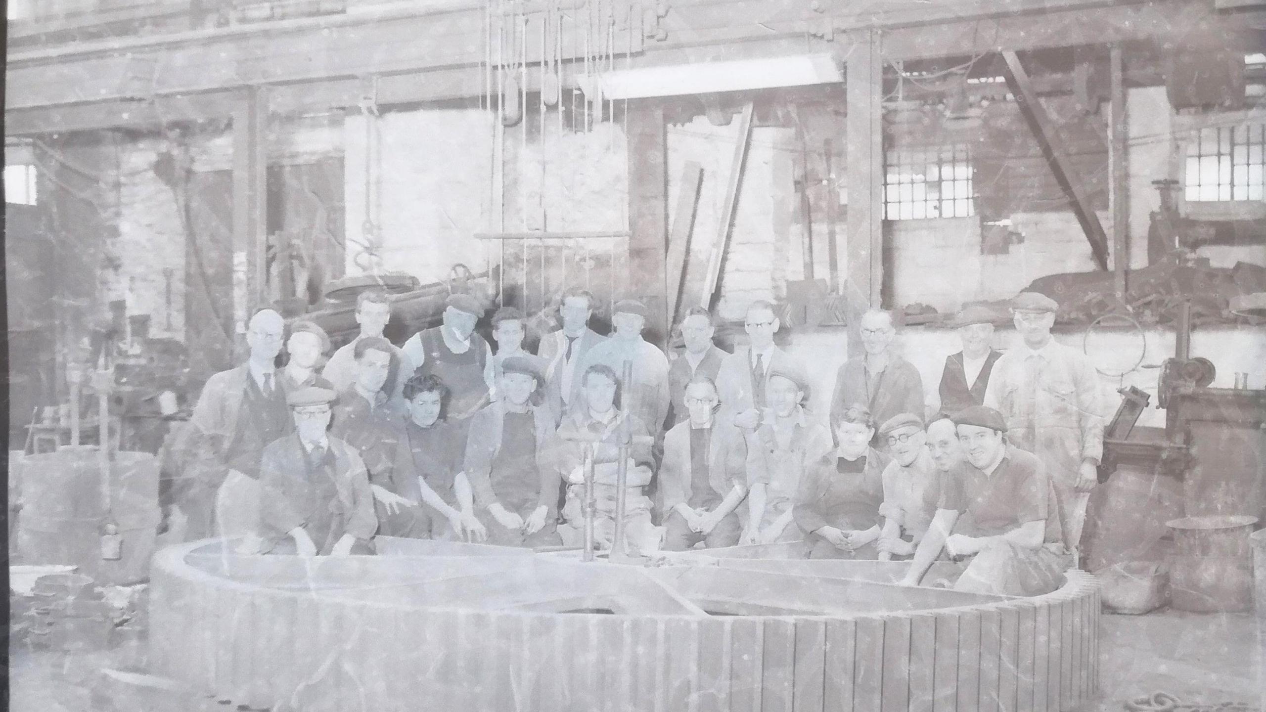 A photographic image shows 23 workers dressed in jackets and caps posing for photographer Henry Wykes at the Bodley Foundry on Commercial Road, Exeter. They are standing and kneeling in front of a huge wheel which is placed on the ground.

