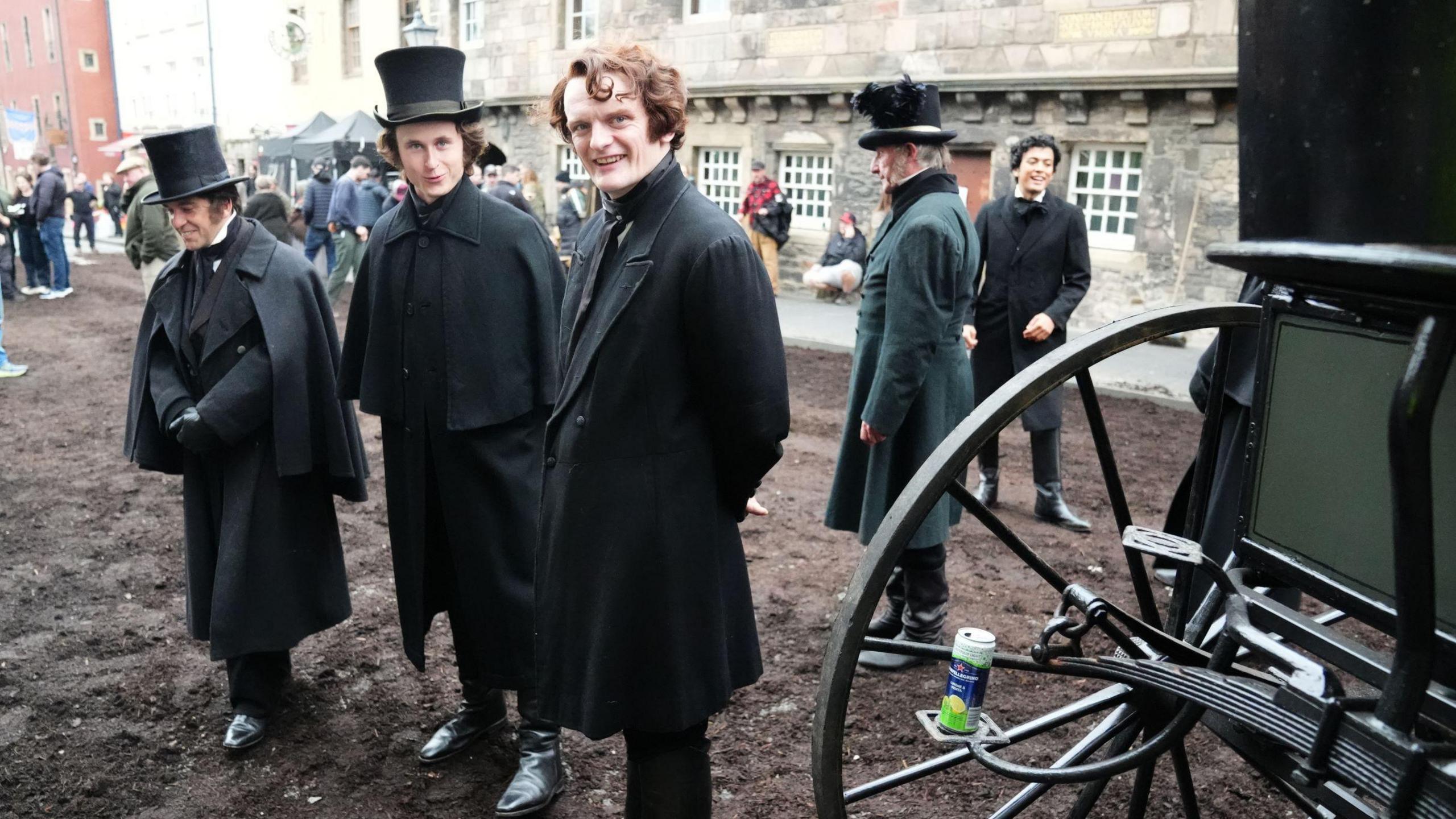 Actors on the set of Frankenstein. Three men are smiling at the camera while dressed in 18th century coats and shoes, with two of the men wearing hats. Other extras mill about in the background. 

The street is all mud and earth. 