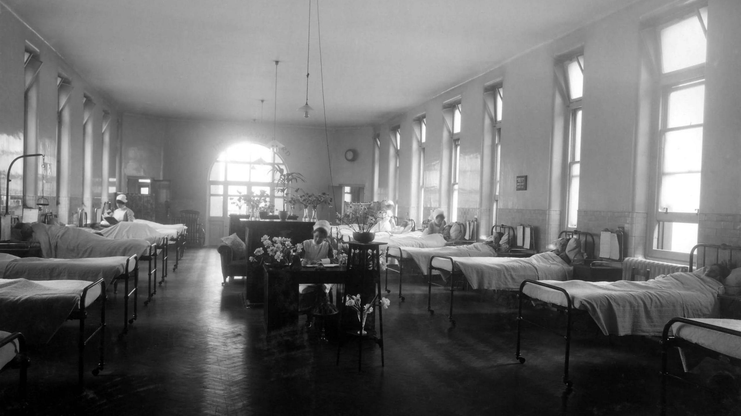 A black and white picture of one of the old wards, with beds lining each side of a large room and a nurse at a table in the middle. There are tall windows and long light fittings and vases of flowers.