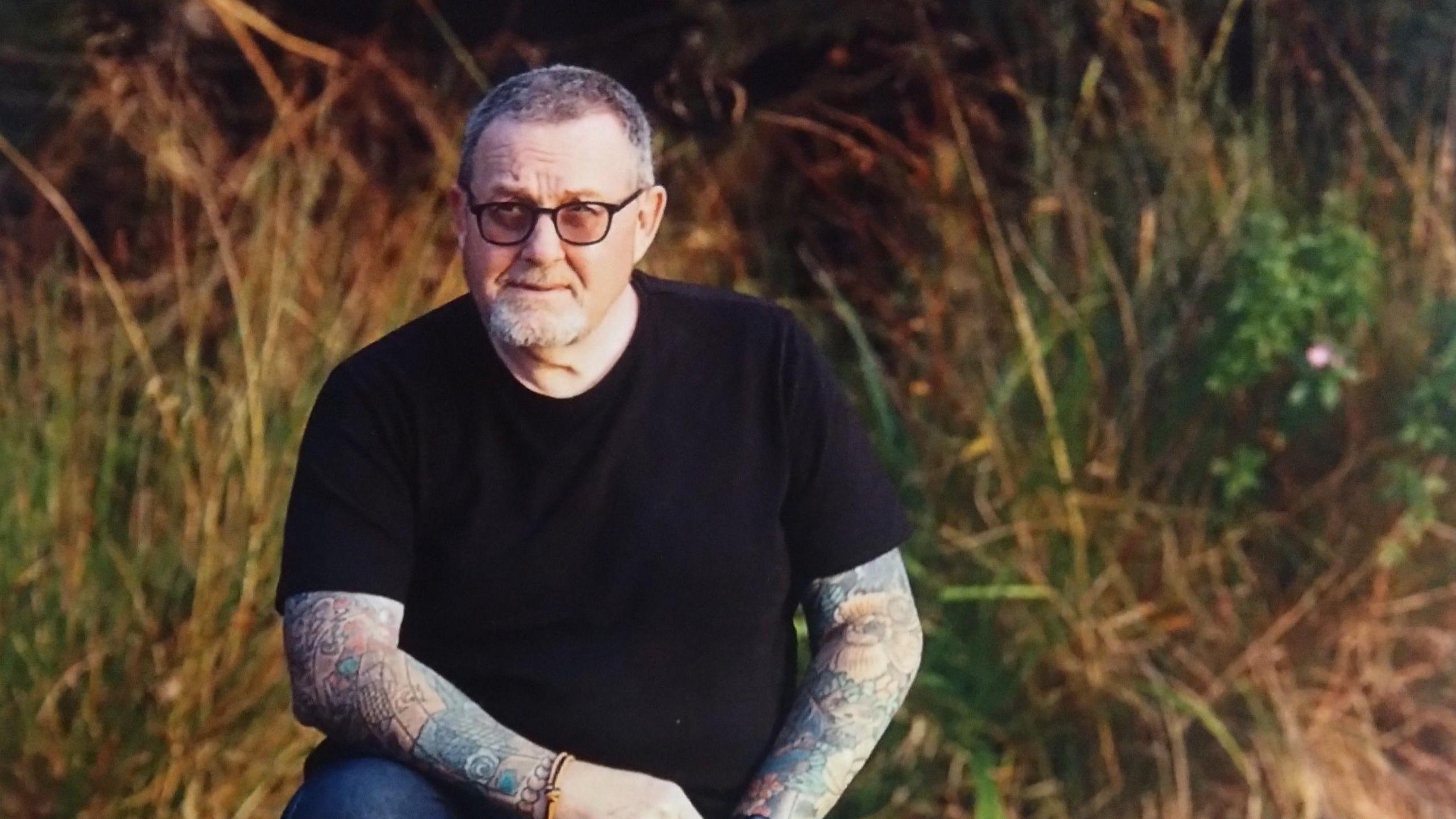 David Spaul has short grey hair and black glasses. He has tattooed arms and is wearing a black T-shirt. He is crouching outside with long grass behind him.