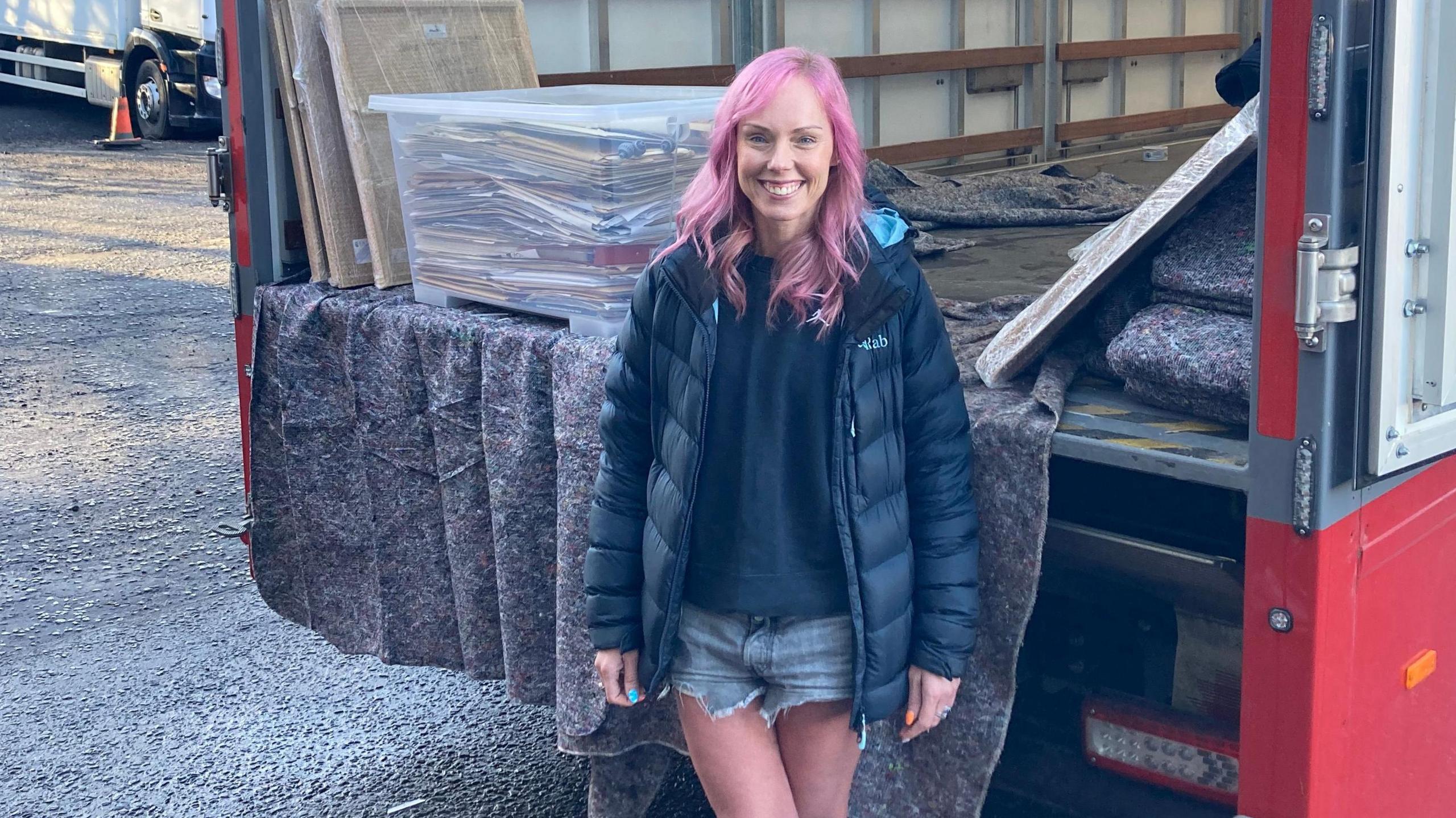 Jane McCormick, in black puffer jacket and denim shorts, is standing in front of the back of an open removals lorry. She is smiling to camera and has pink hair. Part of the archive can be seen behind her.