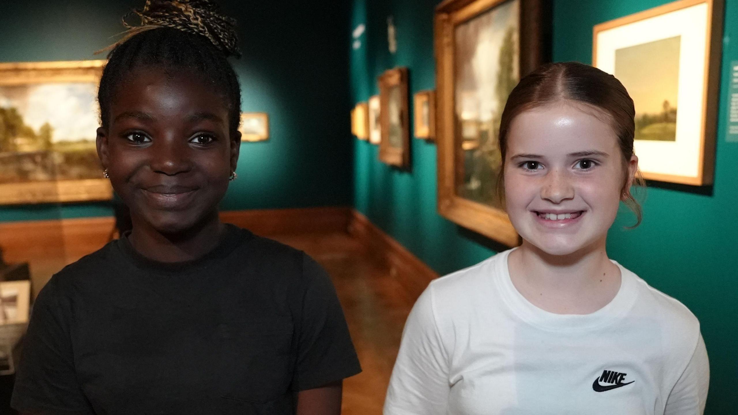 Two young school girls smile at the camera. The one on the left has dark hair that has been tied back and she is wearing a black T-shirt. The girl on the right also has dark hair that has been tied back and is wearing a white T-shirt with a Nike logo on the chest.