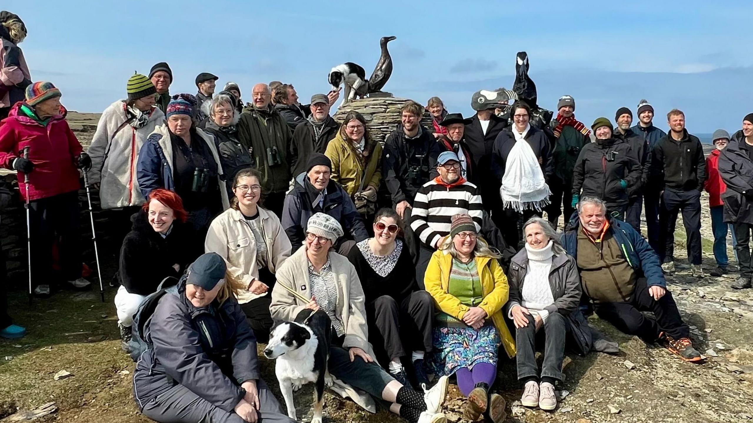 A mixed group of approximately forty people in outdoor clothes are gathered around the Great Auk statue and cairn. Some are sitting on the rocky ground and all are looking at the camera.