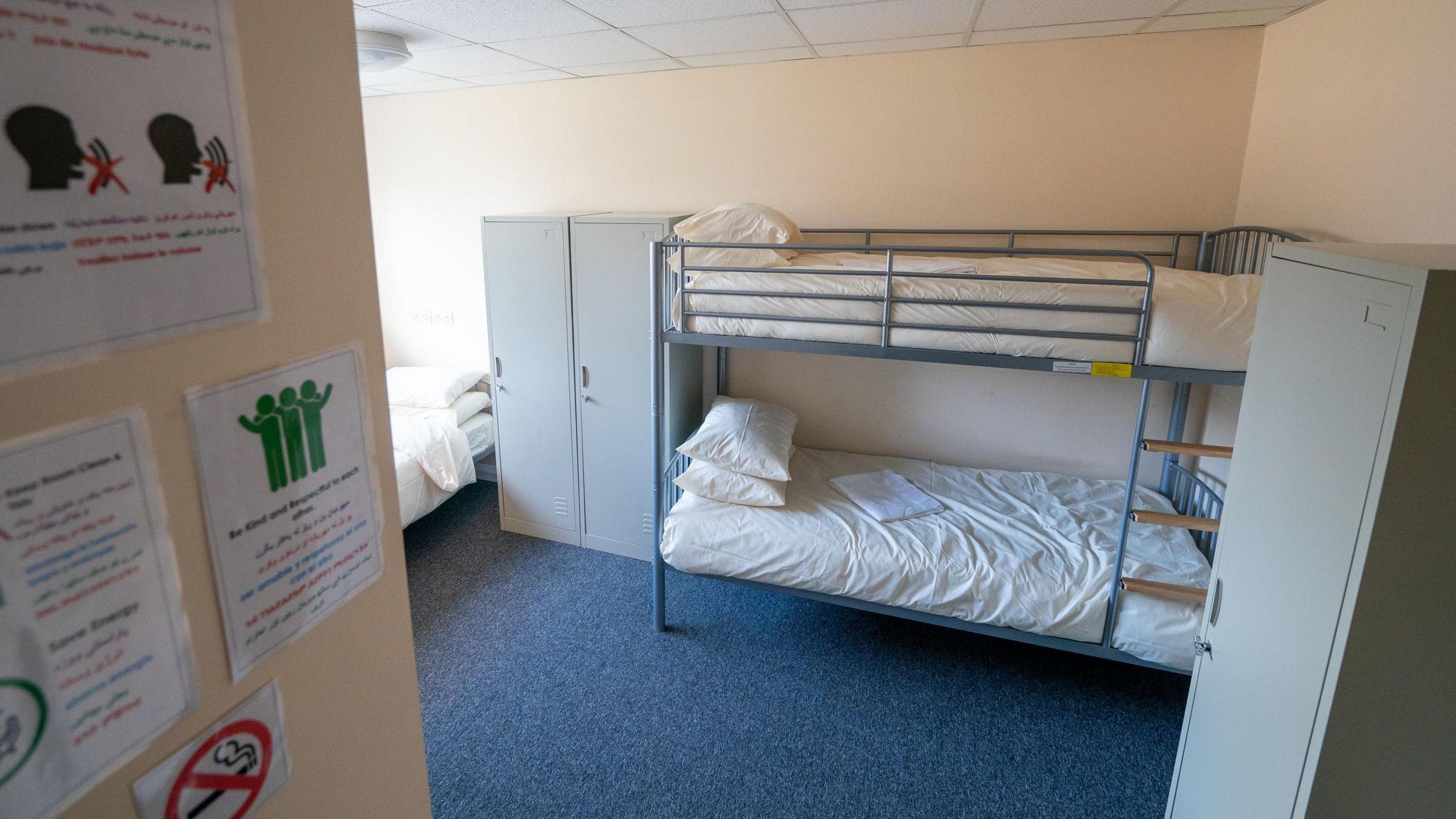 On the right of the image there are bunk beds, that have a metal frame and white pillow and duvet covers. On the left there is another single bed, and a wooden wall with signs on. 