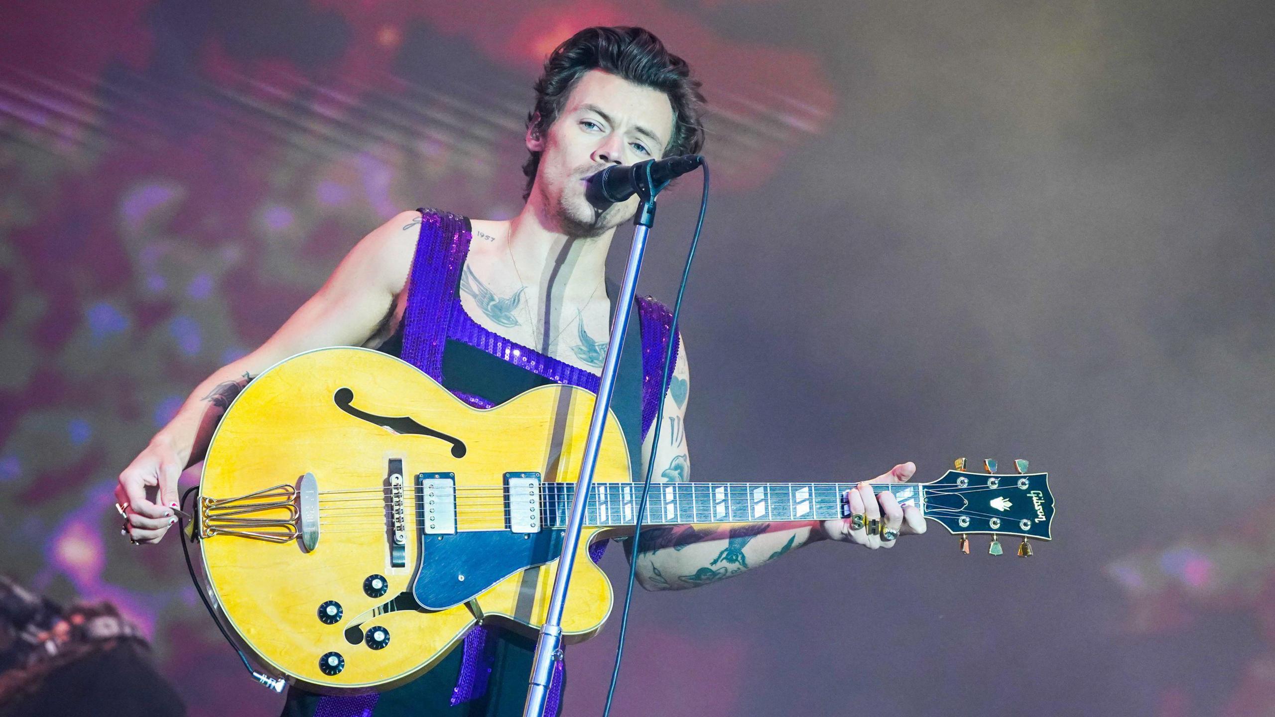 Harry Styles on stage singing into a microphone and holding a bass guitar, wearing a black and purple set of overalls 