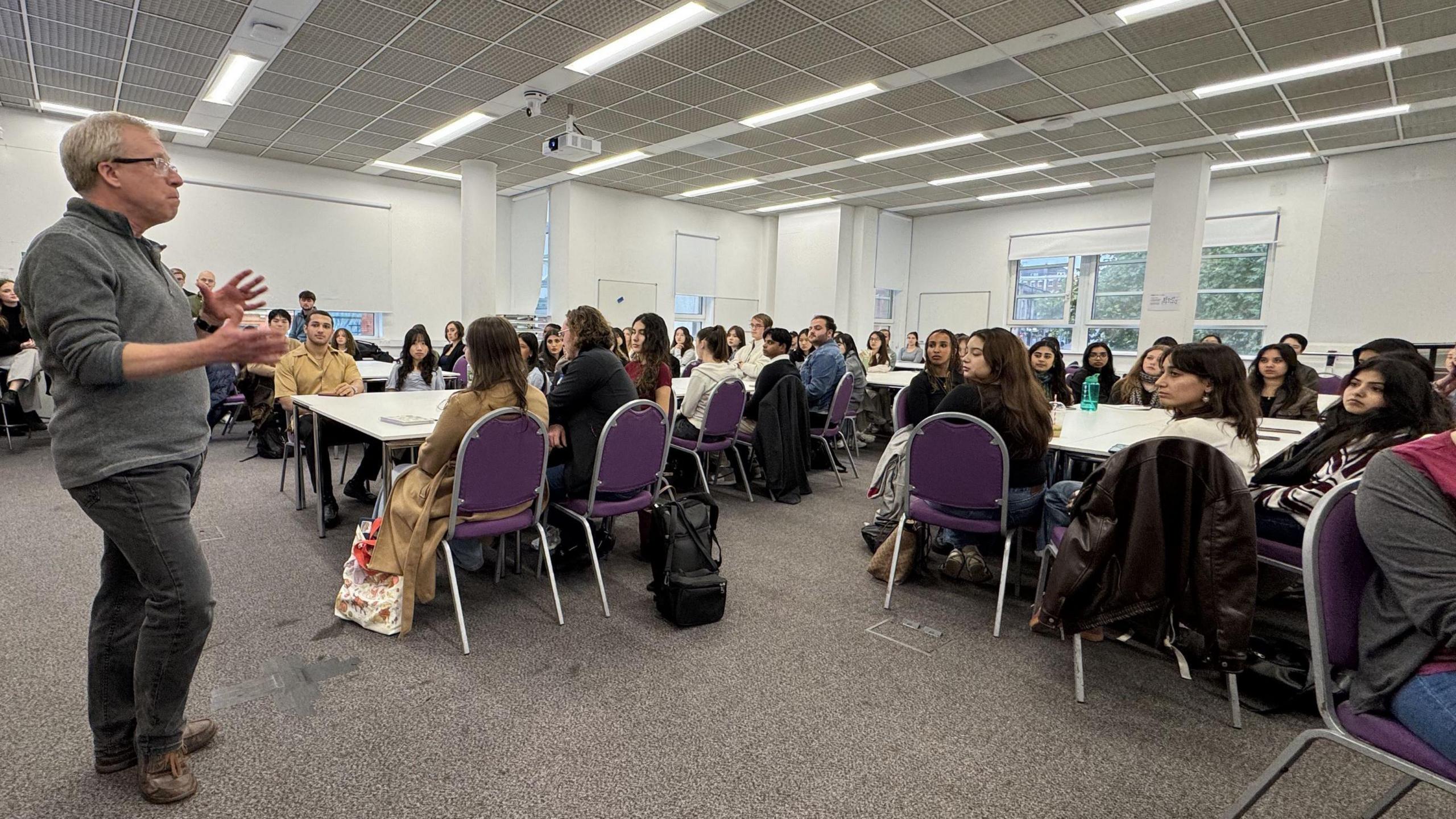 A middle-aged man wearing a light grey jumper, dark trousers and glasses stands as he addresses a room full of law students who are all seated at different tables. The room has a grey carpet, strip lighting overheard and white walls