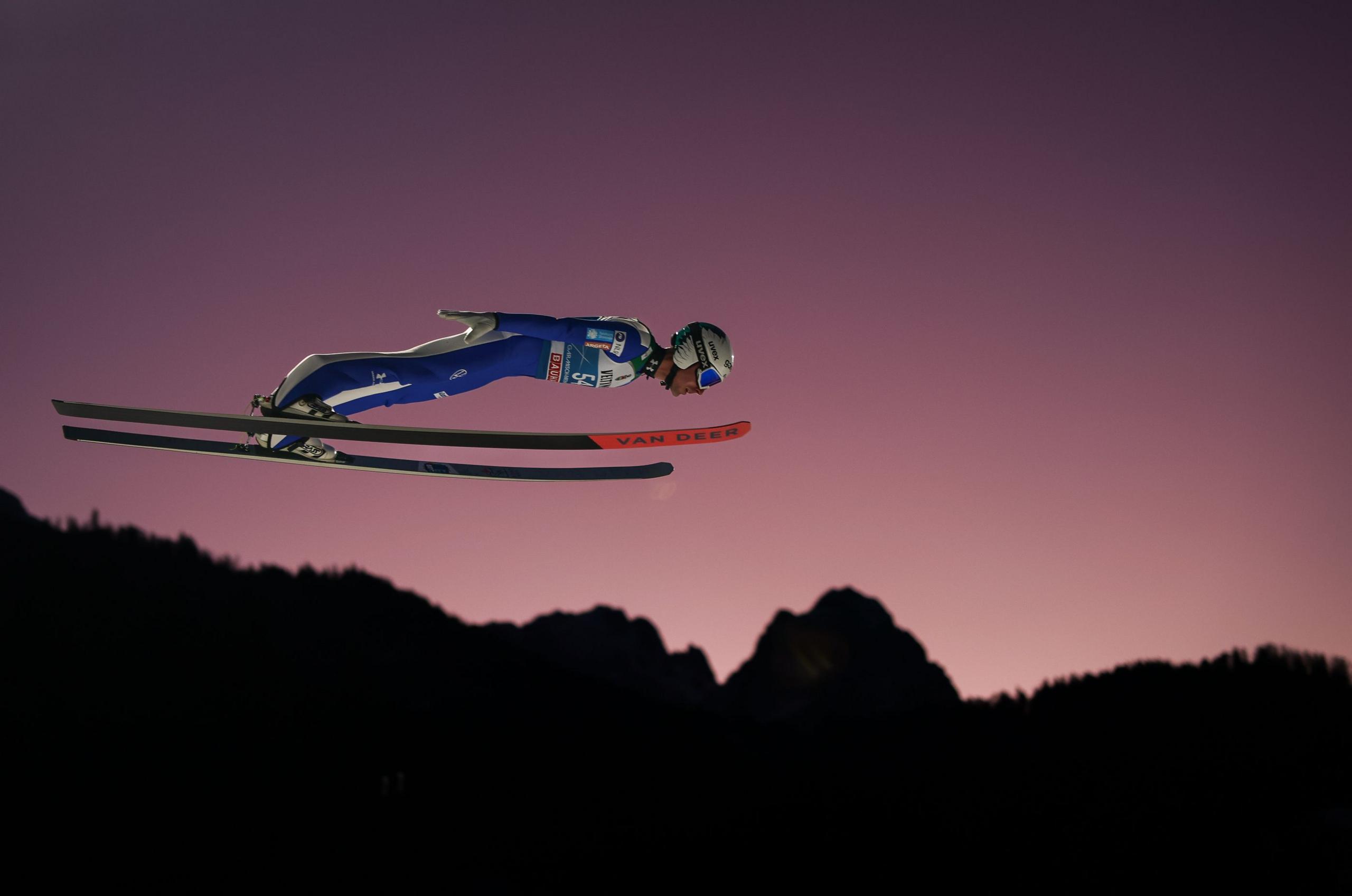 A ski jumper in mid-air during a jump, wearing a blue and white suit with a helmet and goggles. The jumper is in a streamlined position with skis parallel and extended forward. The background shows a silhouette of mountains and trees against a purple-pink twilight sky.