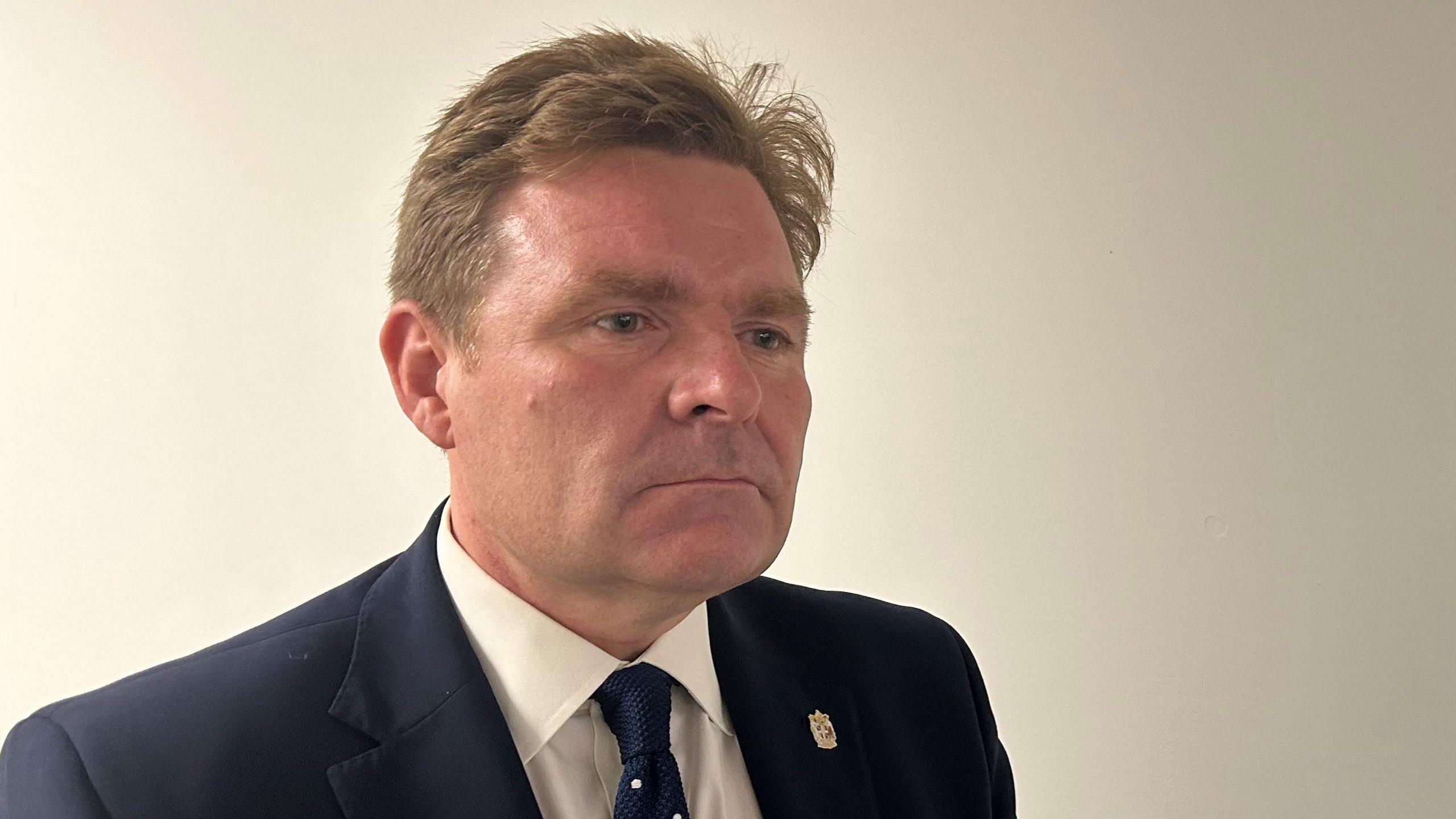 Birmingham City Council leader John Cotton, wearing a navy blue suit with a pin badge on the lapel. He is also wearing a navy coloured tie with white spots.