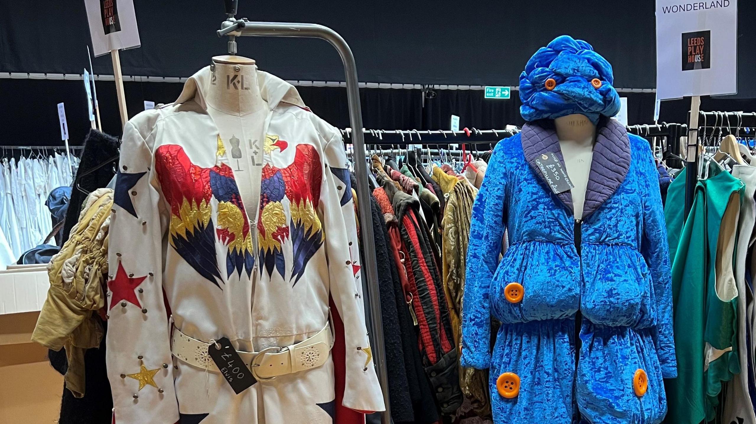 Two costumes on mannequins. To the left is an Elvis-style jumpsuit in white with gold, red and blue detailing. On the right is a blue velvet coat and hat with bulges.