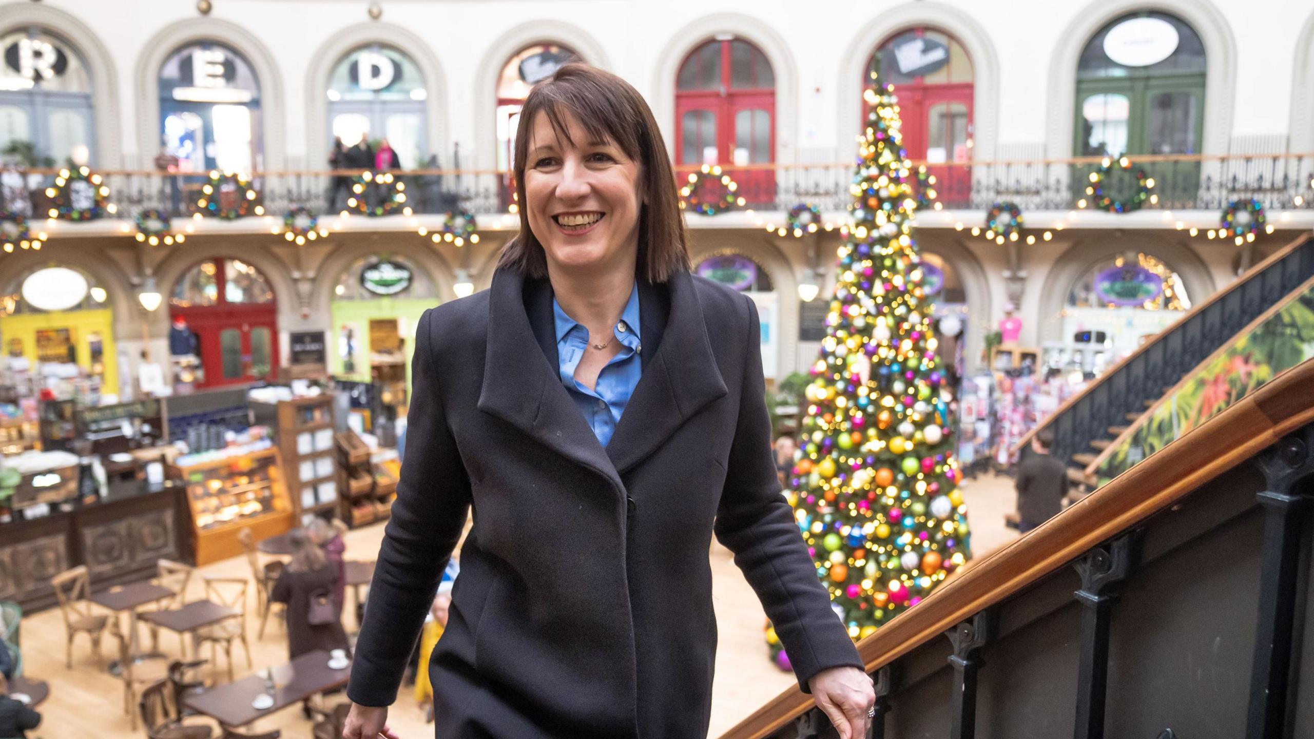 Chancellor Rachel Reeves wearing a dark coloured coat with a blue shirt. She is stood in the Corn Exchange in Leeds with a decorated Christmas tree behind her. 