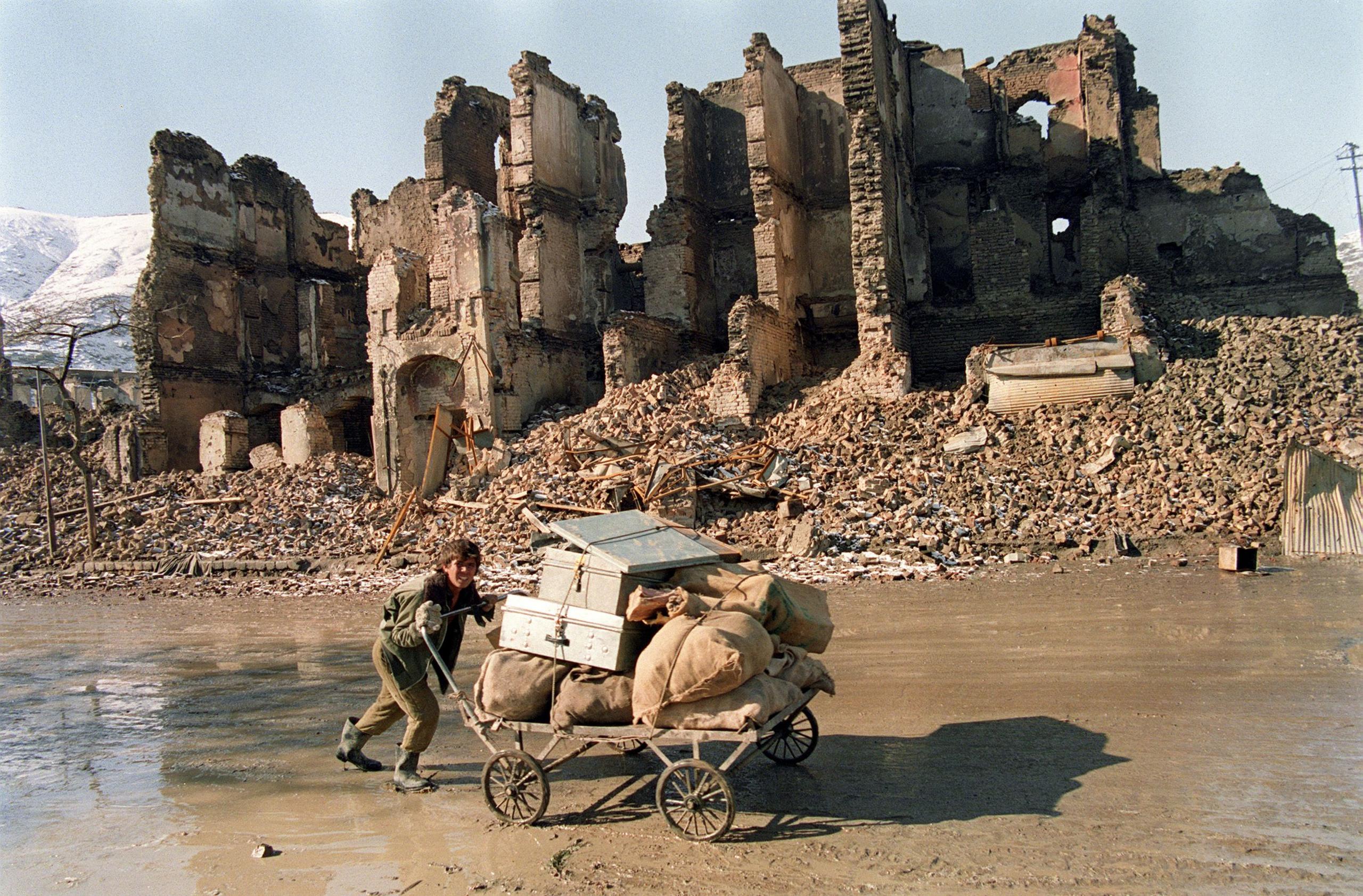 A young Afghan pushes a cart laden with sacks and cases along a muddy street. A destroyed building and rubble can be seen behind him.