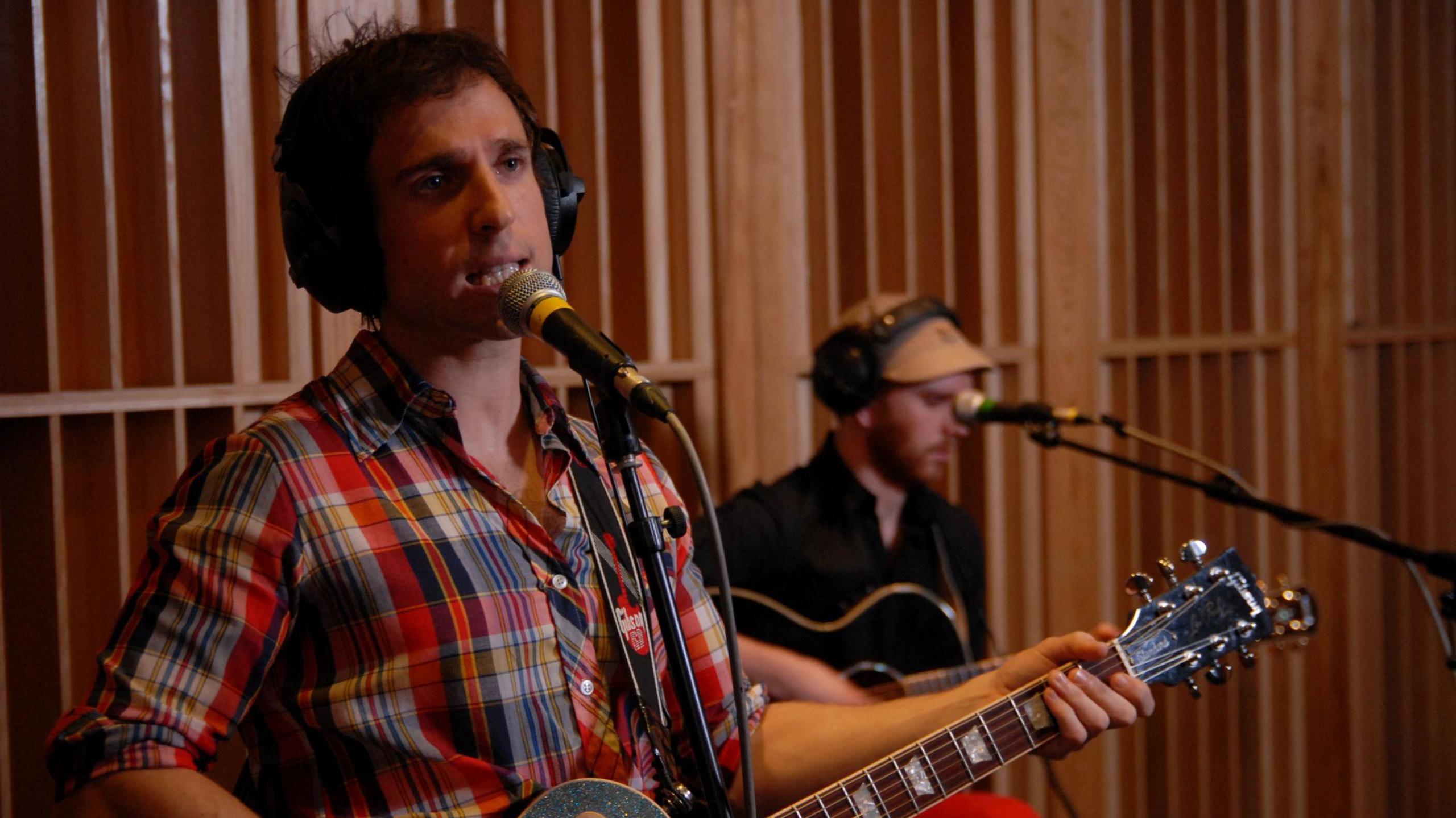 Hoosiers vocalist Irwin Sparkes is wearing a checked shirt and black headphones and singing into a microphone on a stand while holding a guitar. Behind him, Alan Sharland is out of focus, wearing black headphones over a cap and a black shirt. He is also holding a guitar and has a microphone in front of him. Behind them is a wall made of vertical slats of wood.