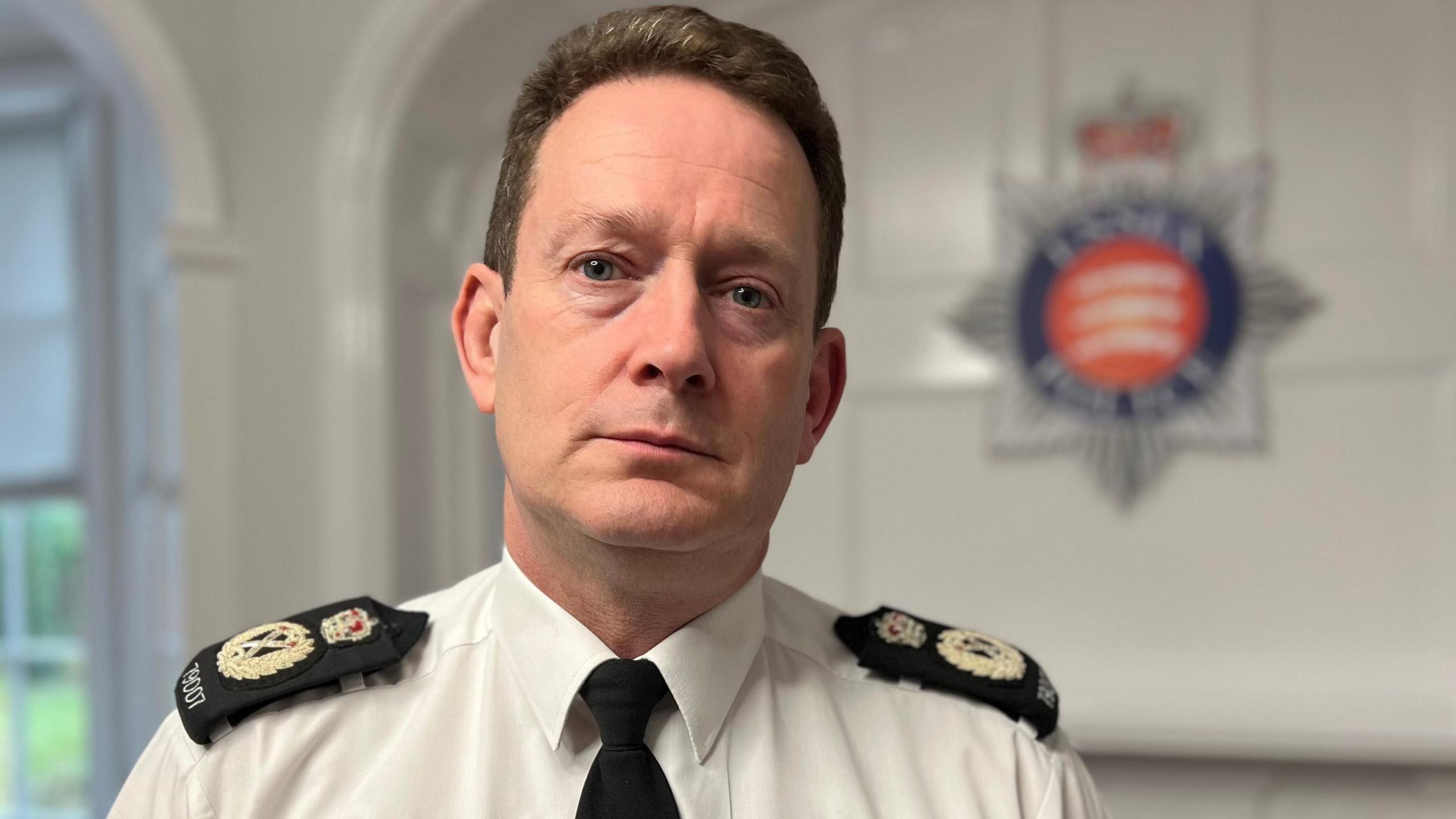 Chief Constable Ben-Julian Harrington, who has short light brown hair. He is wearing a white shirt with blue police lapels and a dark tie. He is sitting in a room with the Essex Police log blurred out on a white wall behind him.