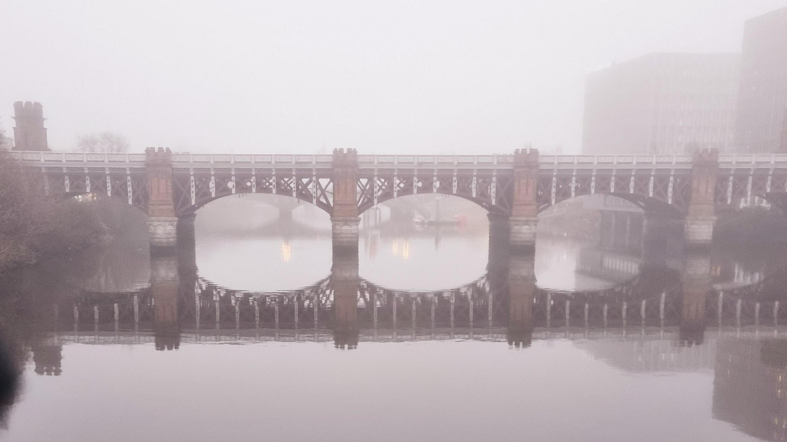 The  Caledonian Railway Bridge over the River Clyde in Glasgow. The bridge has red and white metal connected between red brick turrets. A thick fog is covering the scene and the bridge is reflected in the water below.