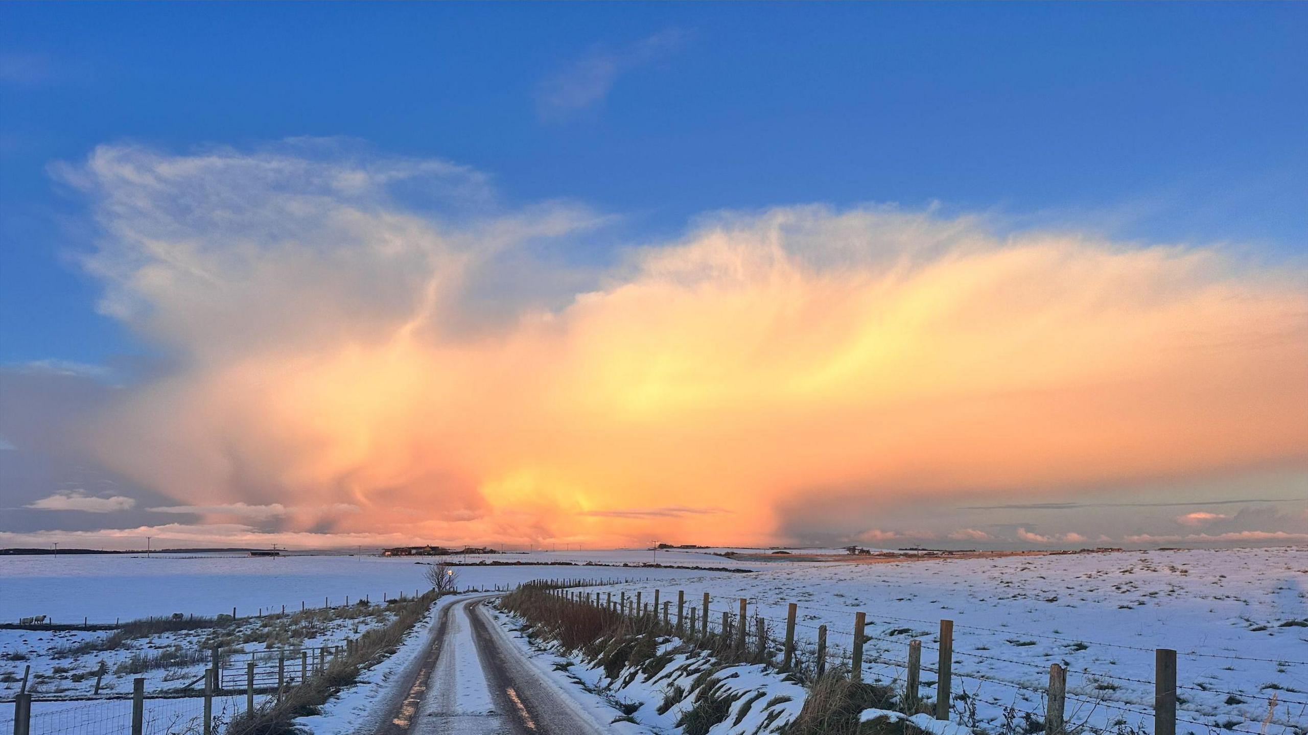 A huge cloud in the sky that is a luminous orange colour. There is a snowy scene underneath of fields, a road and a fence.