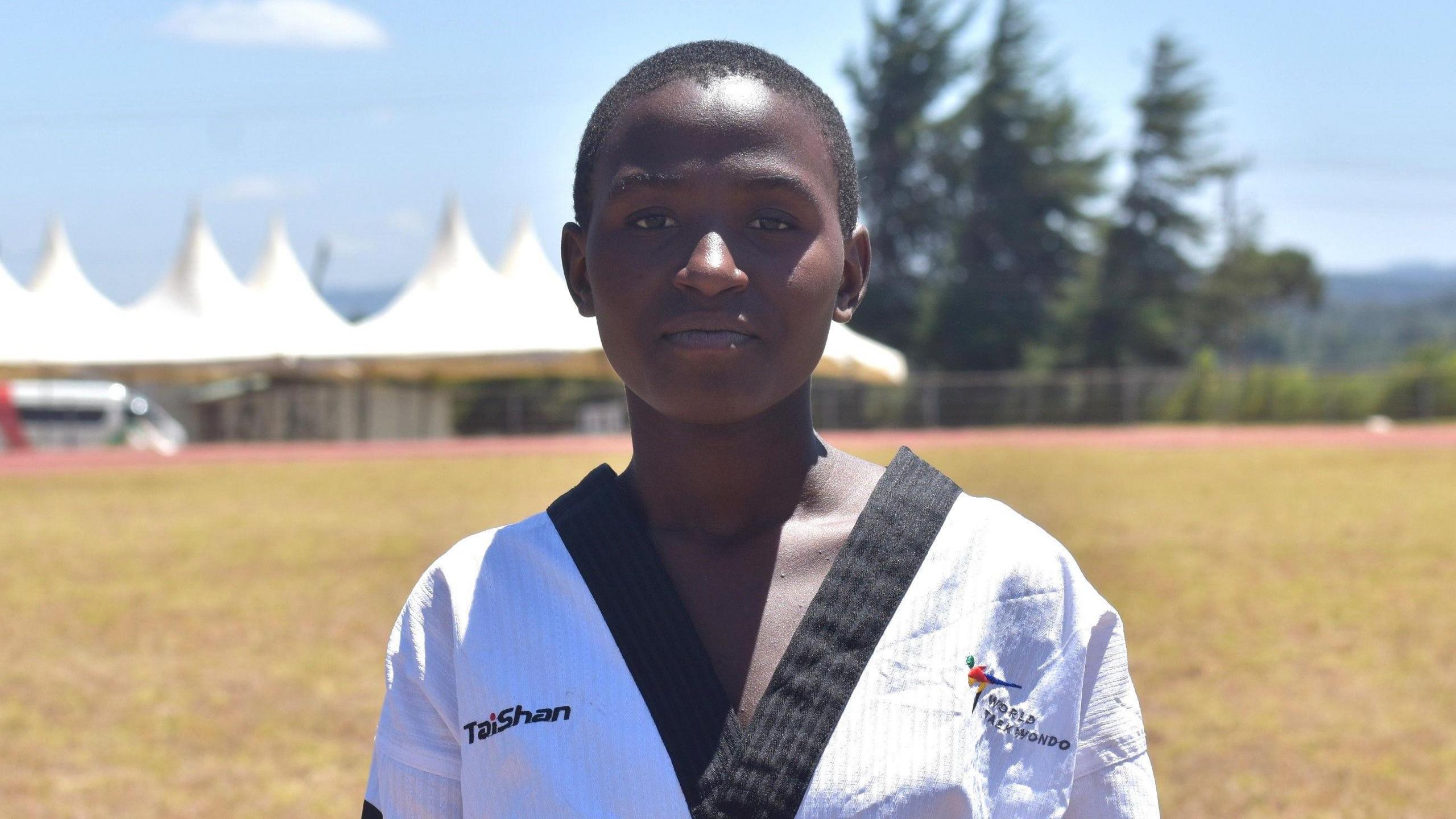 Laurence Namukiza, wearing a white taekwondo outfit with black trim, looks straight at the camera as a large tent and trees are seen out of focus in the background