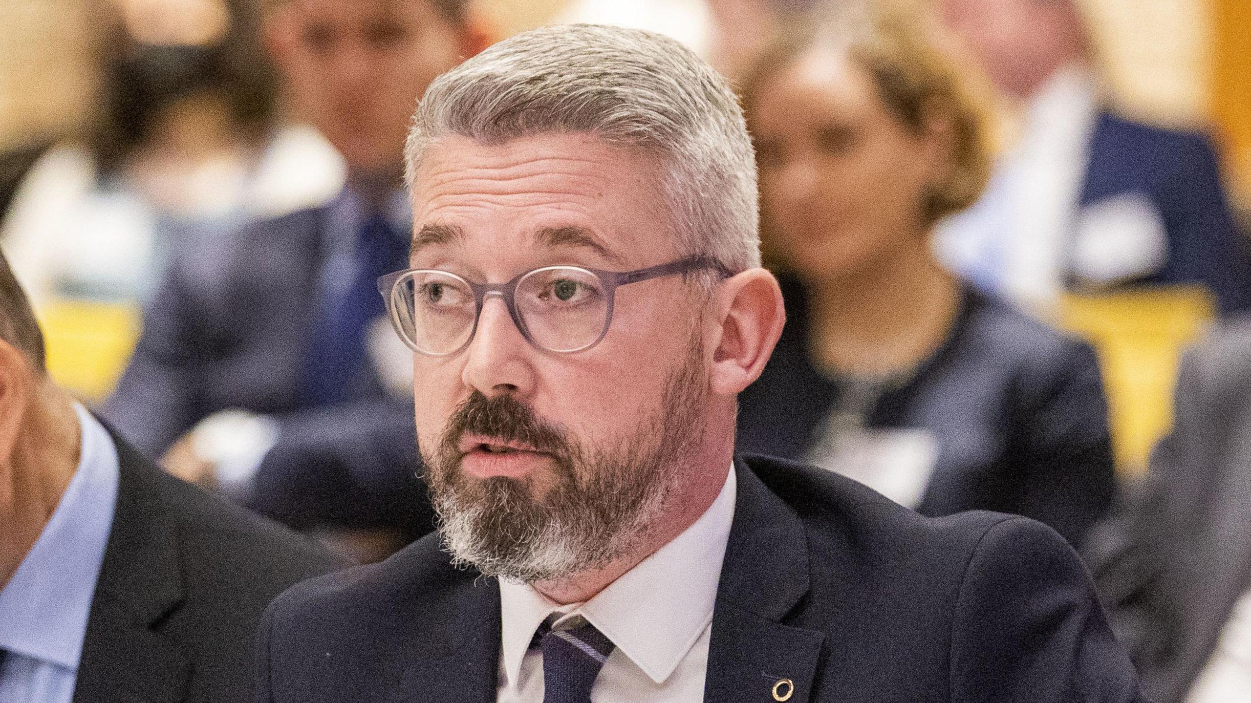 Niall Ó Donnghaile sits in a room surrounded by other faces that are not in focus. He has grey hair and a dark-coloured beard with two grey patches. He also wears grey-rimmed glasses and a black suits with a white shirt and black tie. 