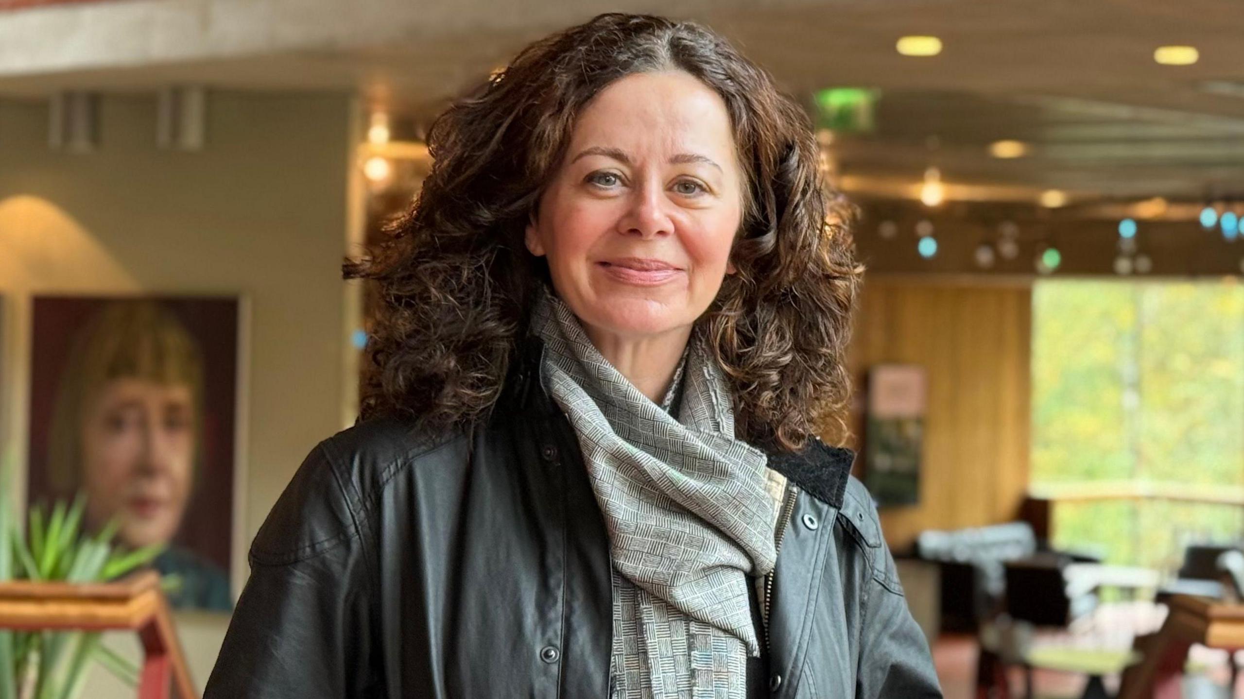 A woman with curly brown hair smiles at the camera. She is wearing a black leather jacket and grey scarf. There is a cafe behind her. There are large photos on the wall.