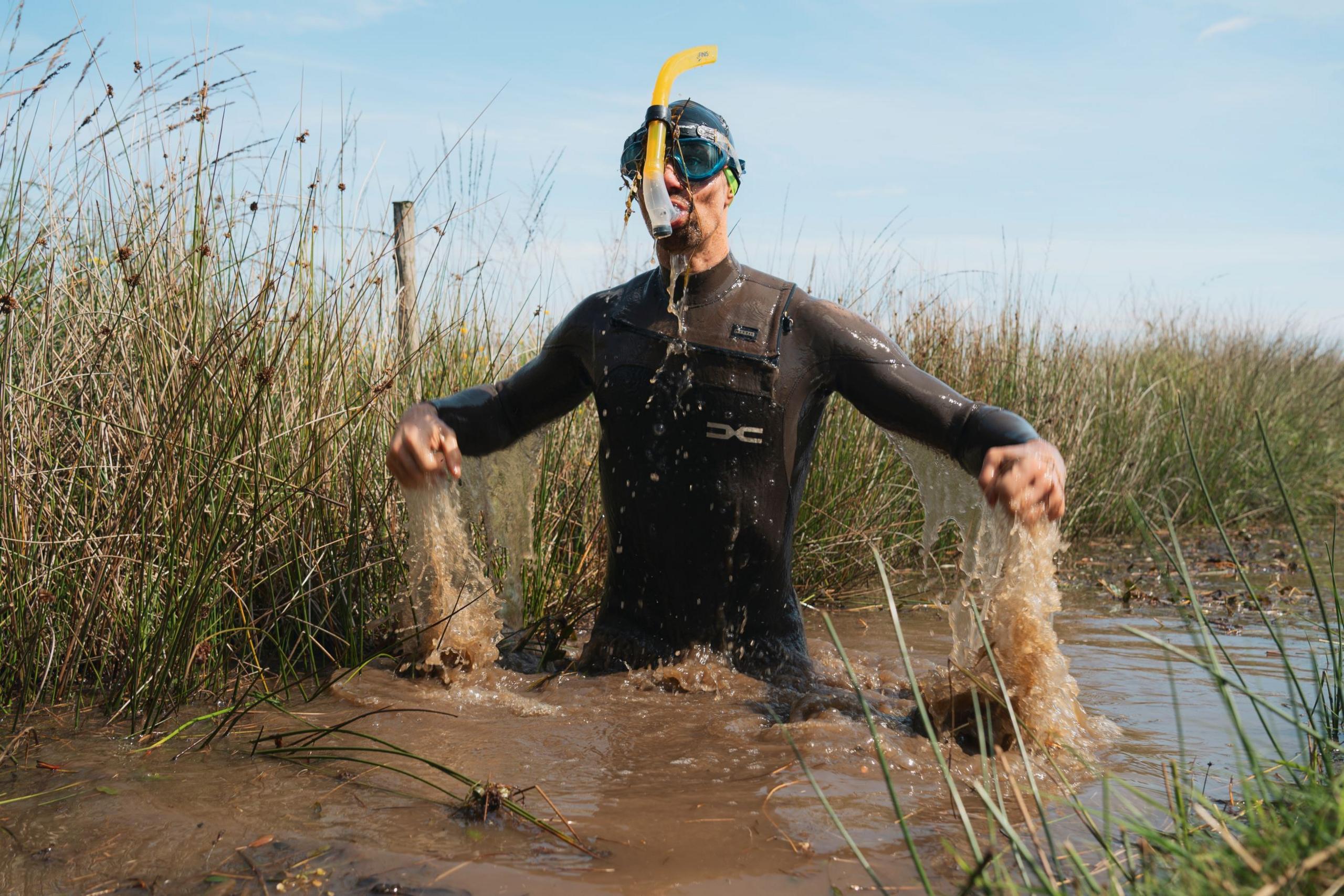 Neil Rutter pictured rising from the bog water, which appears muddy. He wears a long-sleeved wet suit, a hair cap and a snorkel. Blue skies can be seen behind him and wild grass lines the edge of the water. 