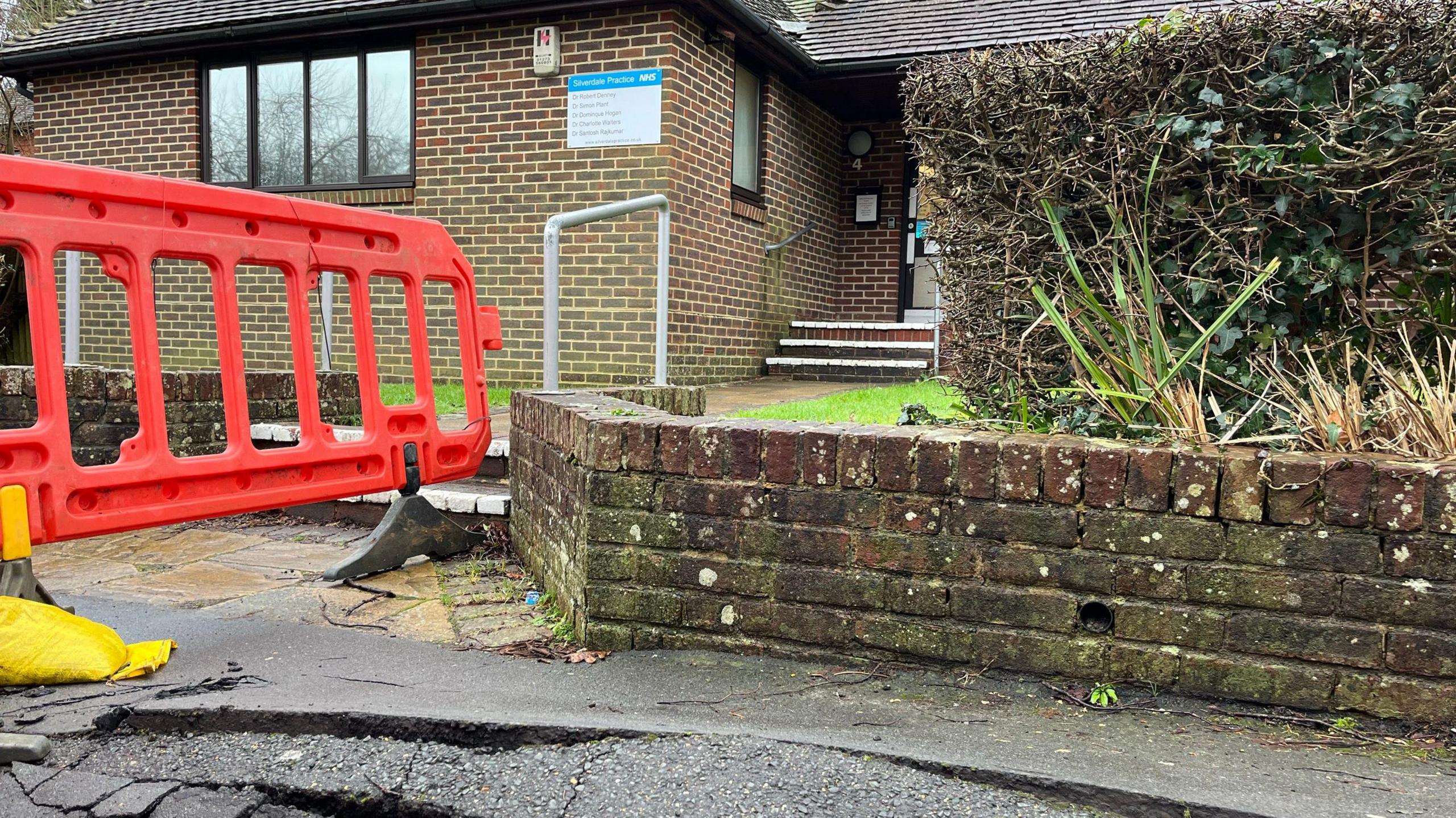 Silverdale Surgery, a small surgery with one floor. It has a small front garden which leads out to the pavement which was said to have been damaged by tankers dealing with the situation. The pavement is raised and uneven and there is orange plastic fencing cordoning the area off