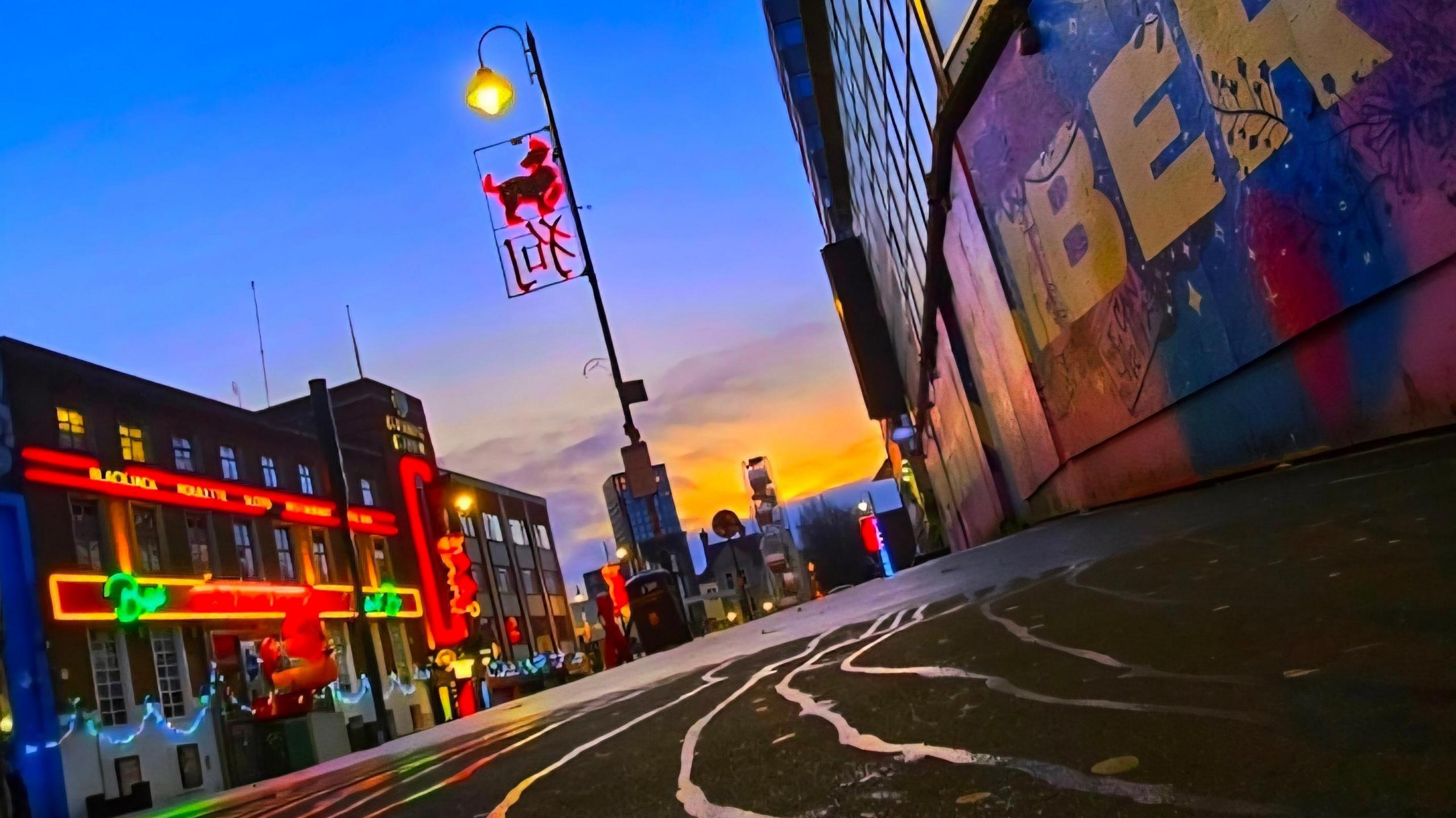 An orange sunrise is captured behind restaurants lit on the exterior with neon signs. A lamppost can be seen in the foreground with a Chinese symbol, also lit in neon