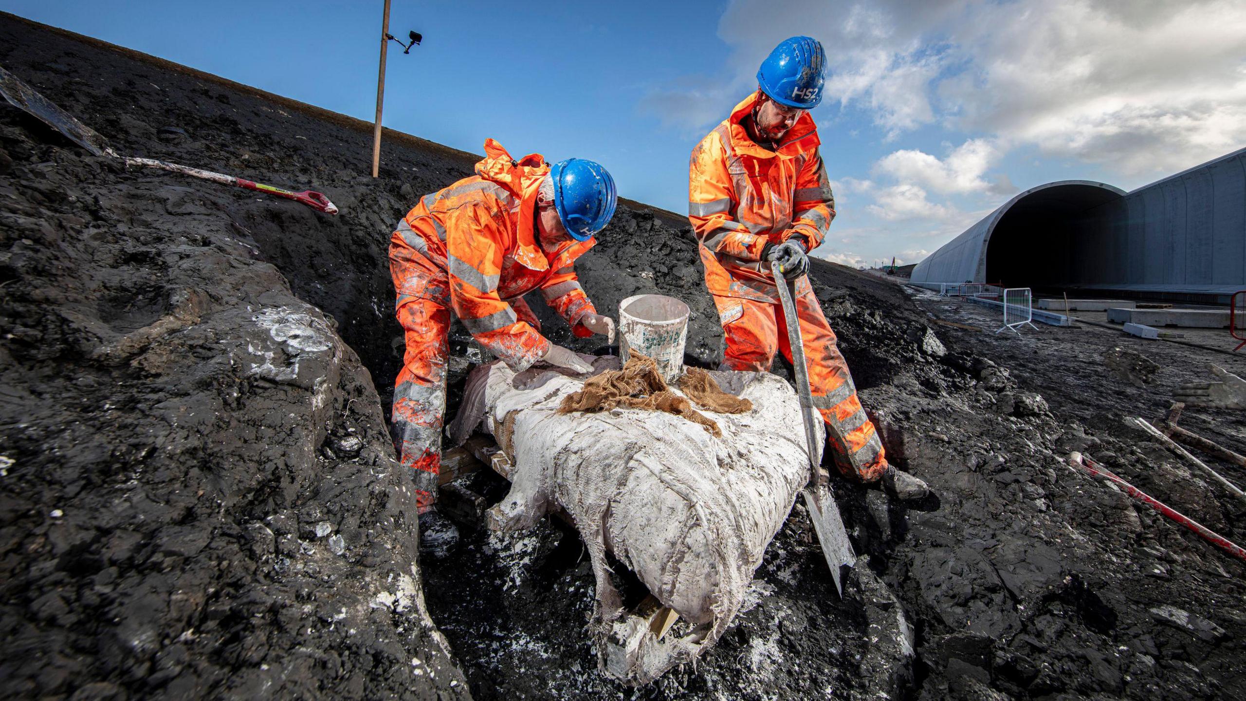 Two men in orange hi-vis clothing excavate a large rock from some dirt.