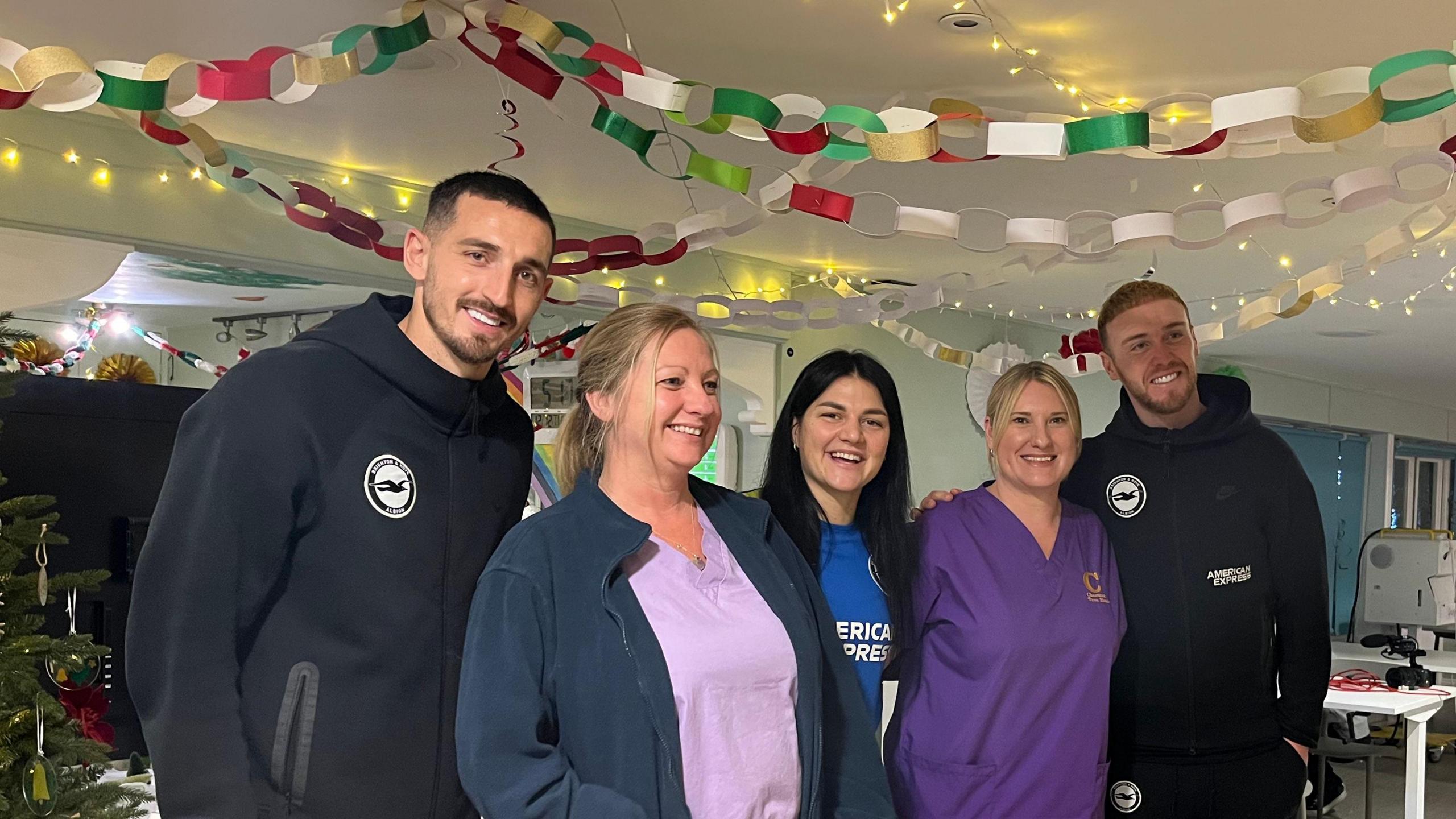 Captain Lewis Dunk, goalkeeper Jason Steele, and women's midfielder Dejana Stefanovic smile for the camera with two hospice staff wearing purple and pink scrubs under Christmas decorations from the ceiling and a Christmas tree to the side.