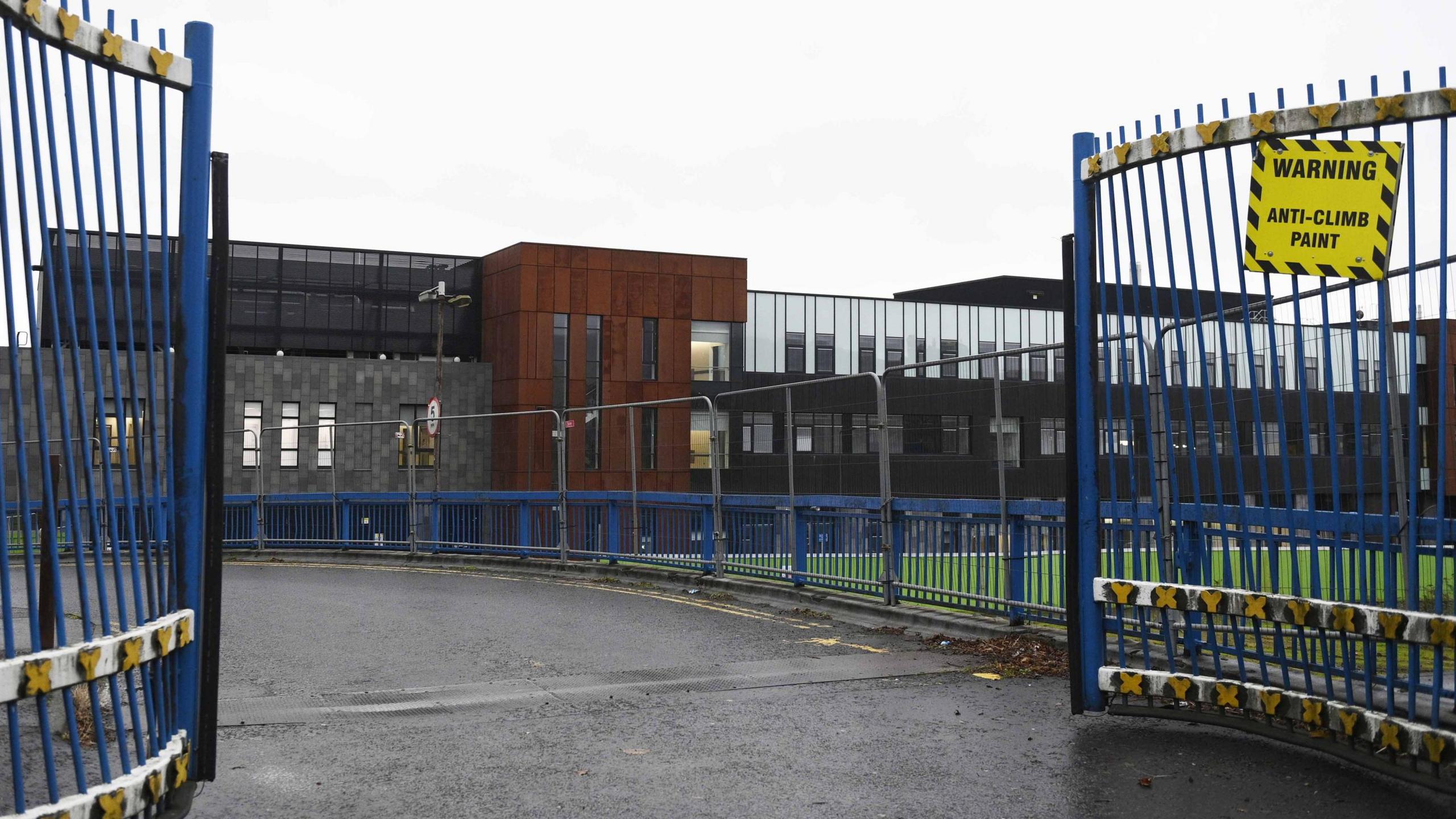 A modern building built in a series of grey and black rectangle shapes with the front a brown cube shape. The blue and white gates at the front are opened. A yellow sign reads, warning, anti-climb paint. The sky is very cloud and it's been raining.