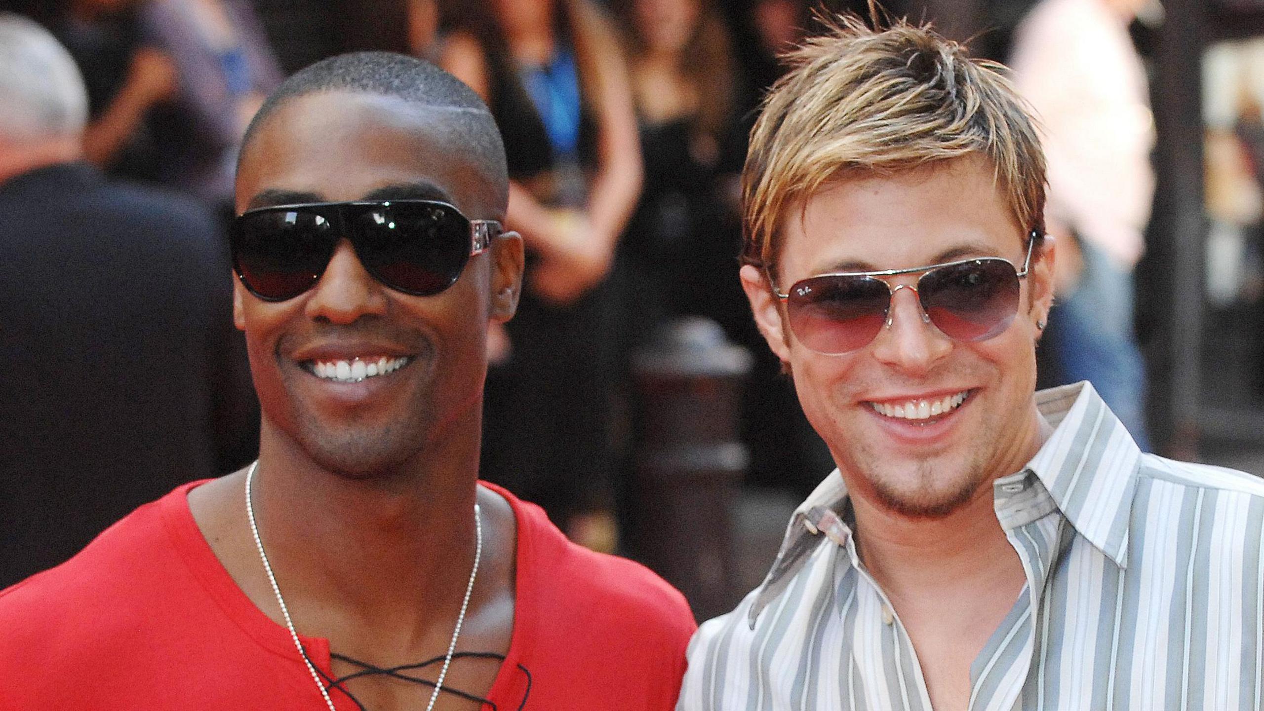 Simon and Duncan stand smiling, each in a pair of sunglasses with a blurred crowd behind then. Simon wears a red T-shirt with a black lace, Duncan wears a white, grey and peach vertically striped shirt. Both have facial hair. 