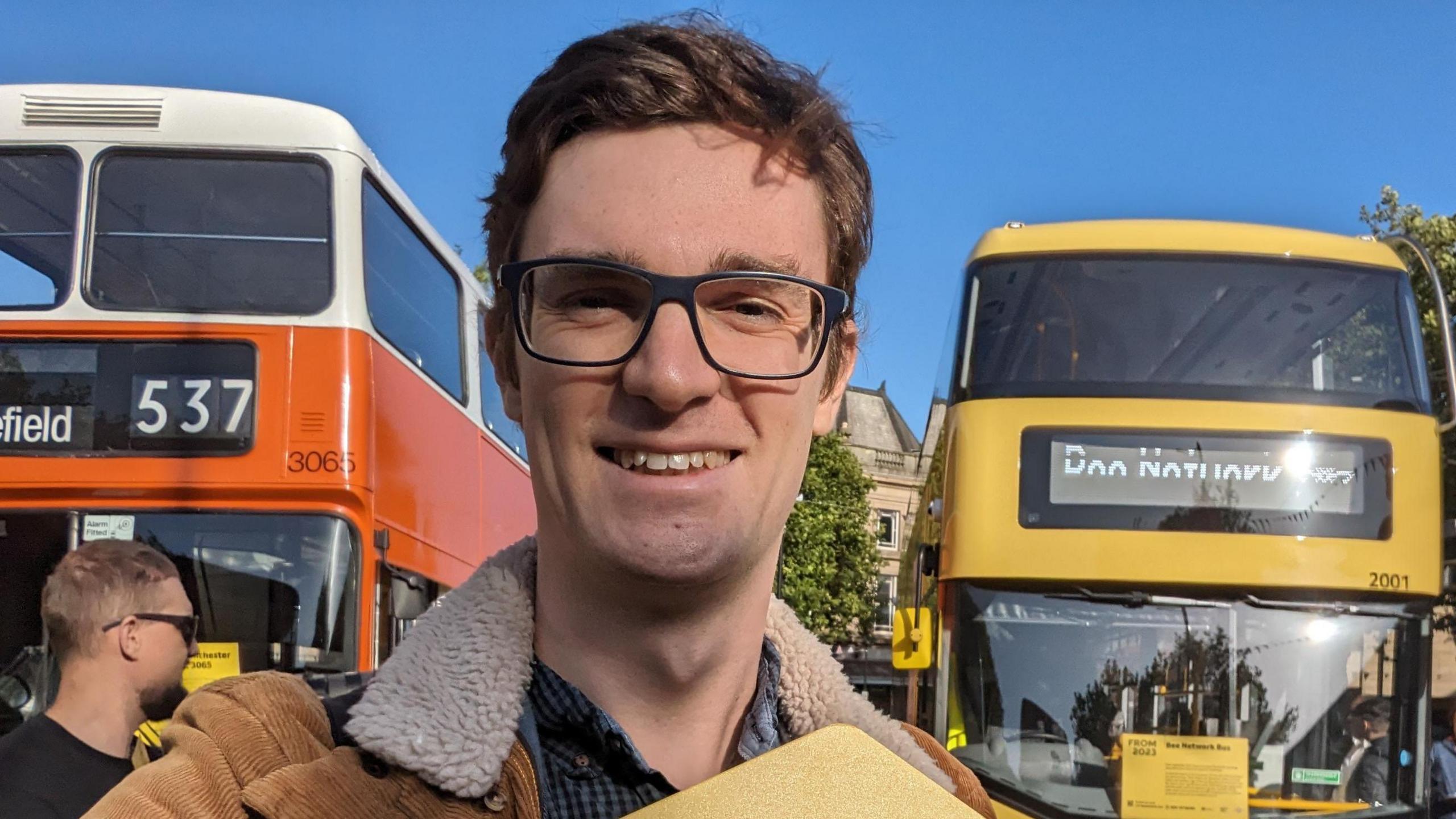 Smiling man with dark brown hair and dark framed glasses, wearing a coat, stands in front of a bright orange bus and a bright yellow bus