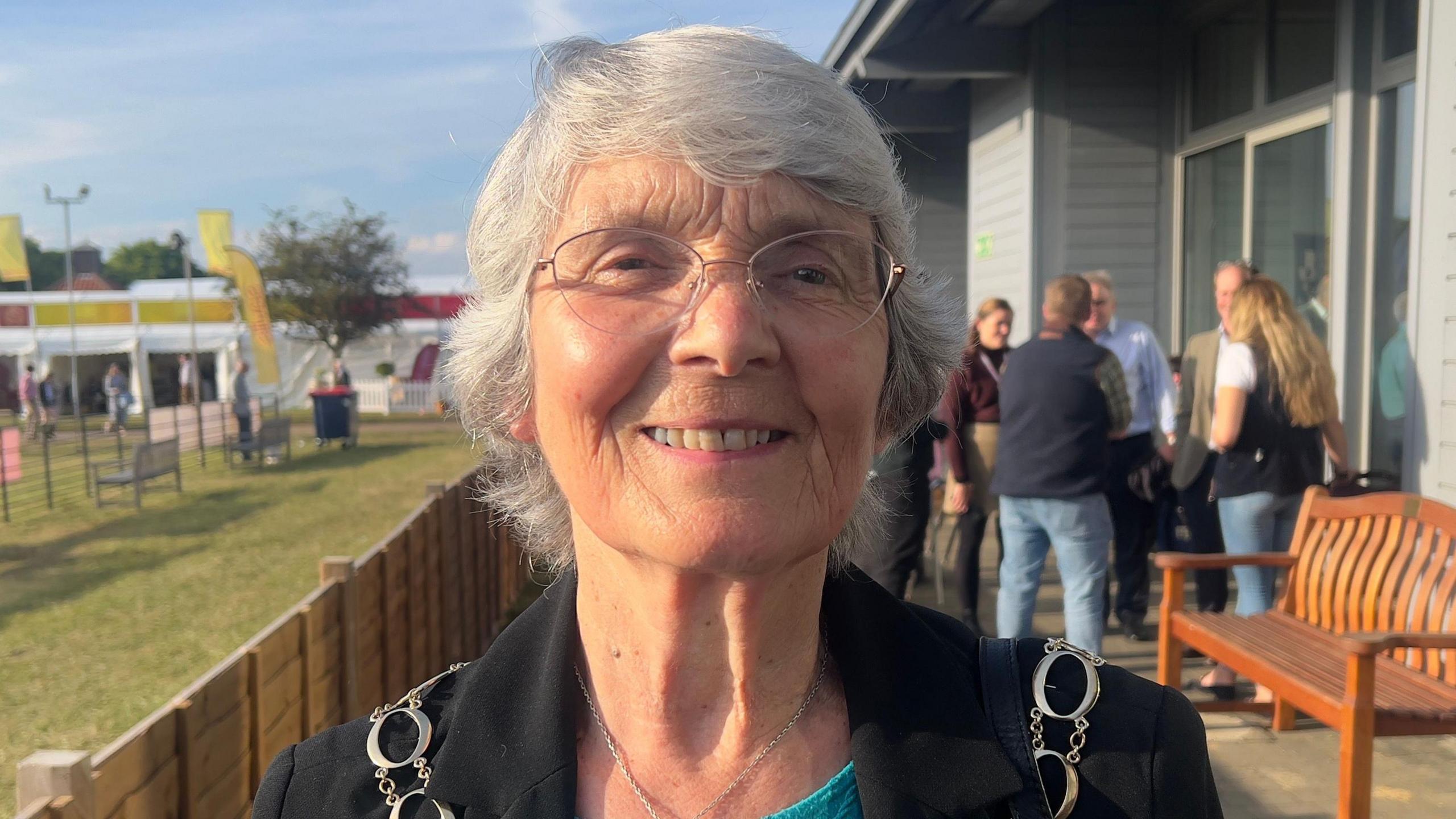 Lesley Hill is looking towards the camera smiling. She has short grey hair and is wearing glasses. She has a silver chain around her, and is wearing a black blazer and a blue T-shirt.