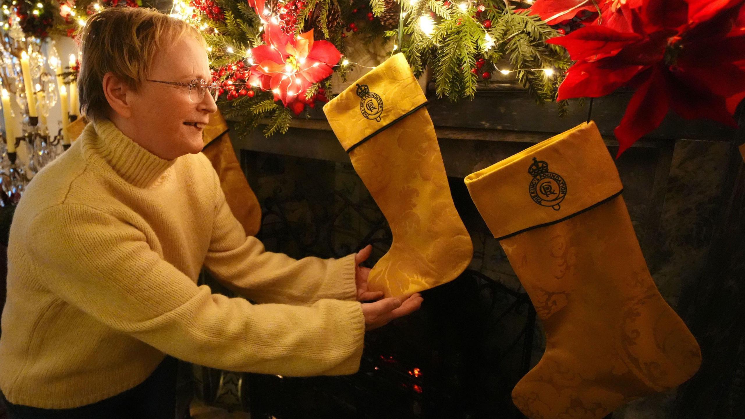 Andrea Middlemiss has short blonde hair and is wearing glasses and a yellow jumper. She is holding one of three stockings fixed to a fireplace decorated for Christmas. The yellow satin damask material has been made into a stocking with black edging and a crest.