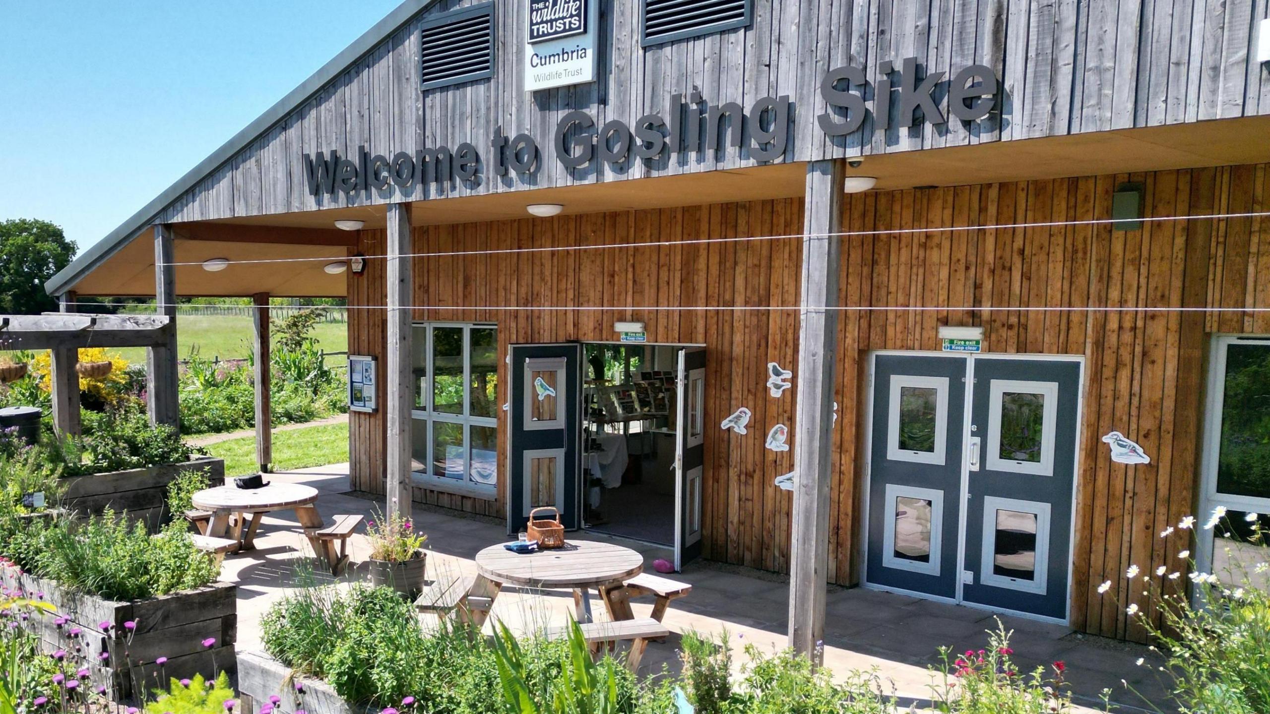 Gosling Sike has a one-storey wooden building with a seating area outside overlooking different flowers and plants. Welcome to Gosling Sike is written above the entrance doors. 