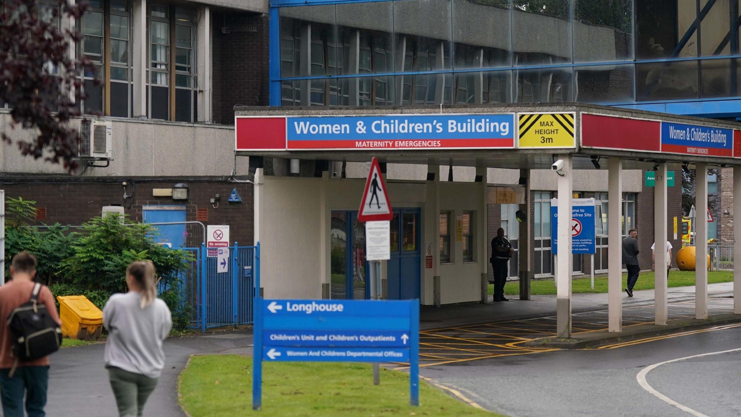 Entrance of hospital - sign reads Women & Children's Building - a man with a rucksack and a woman in a grey sweater are walking towards the door 